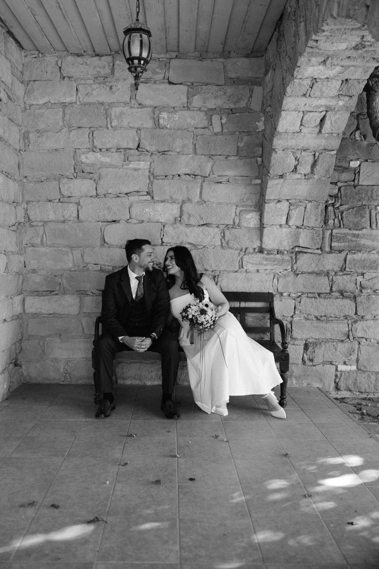 A romantic black and white photo of newlyweds sitting on a bench against a rustic stone wall.