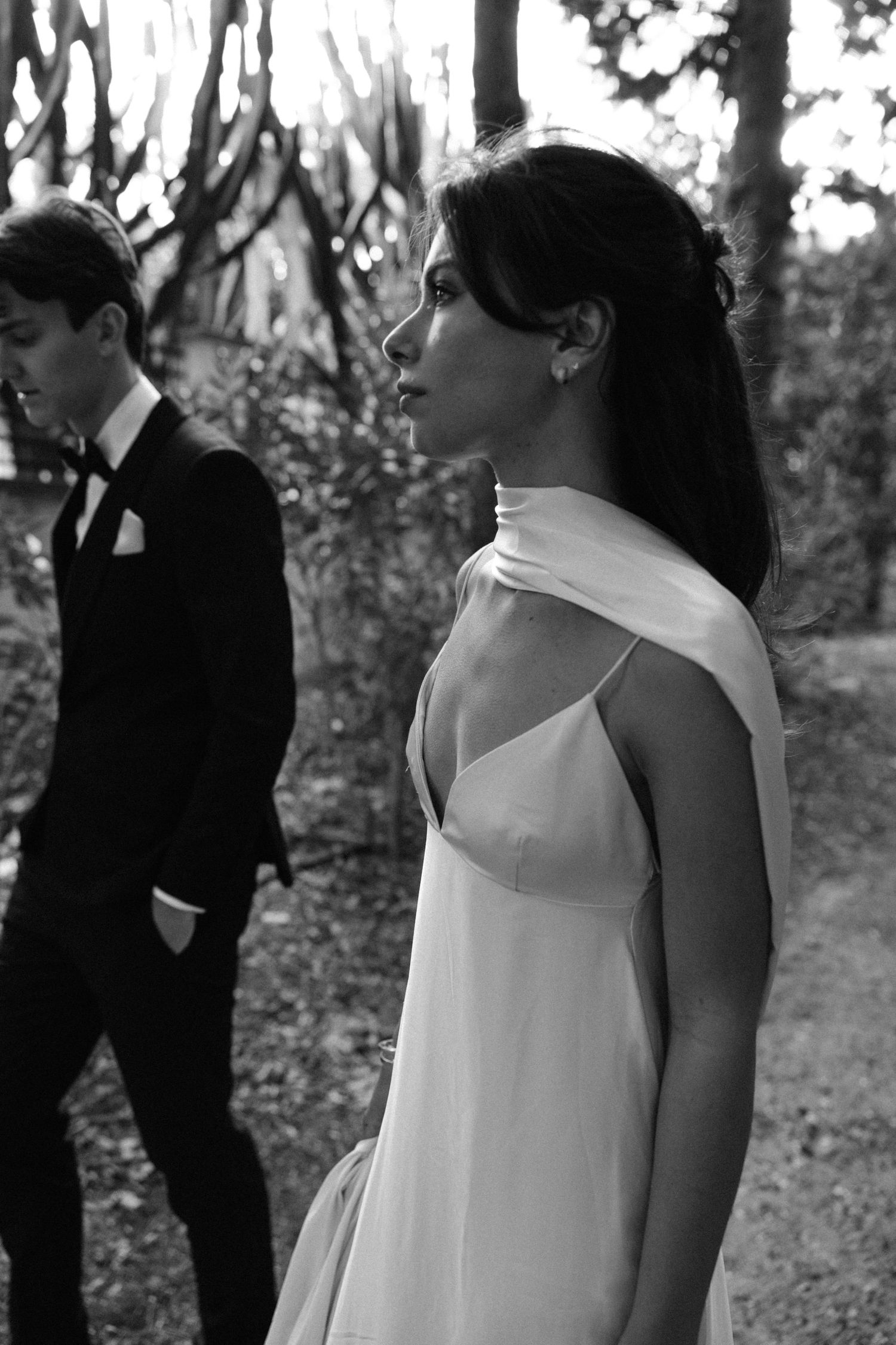 A black and white wedding photo showing a bride in an elegant white dress and groom in a tuxedo outdoors.