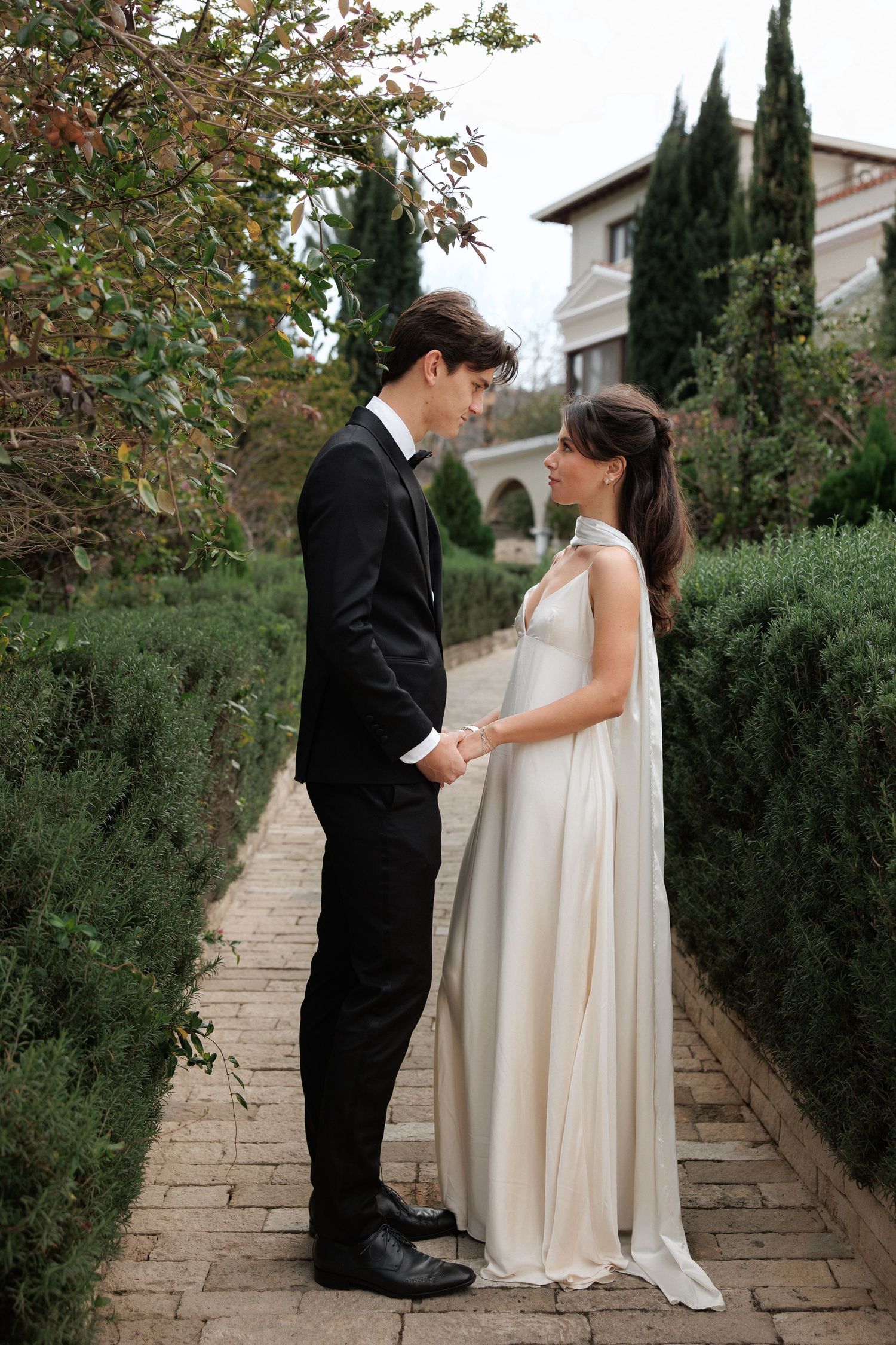 Garden path portrait of couple in wedding attire standing close together among cypress trees.