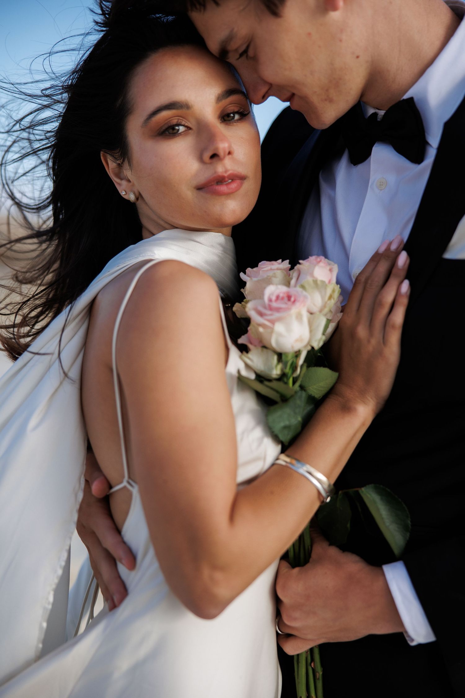 Romantic wedding portrait of couple embracing outdoors with pink roses and flowing hair in the wind.