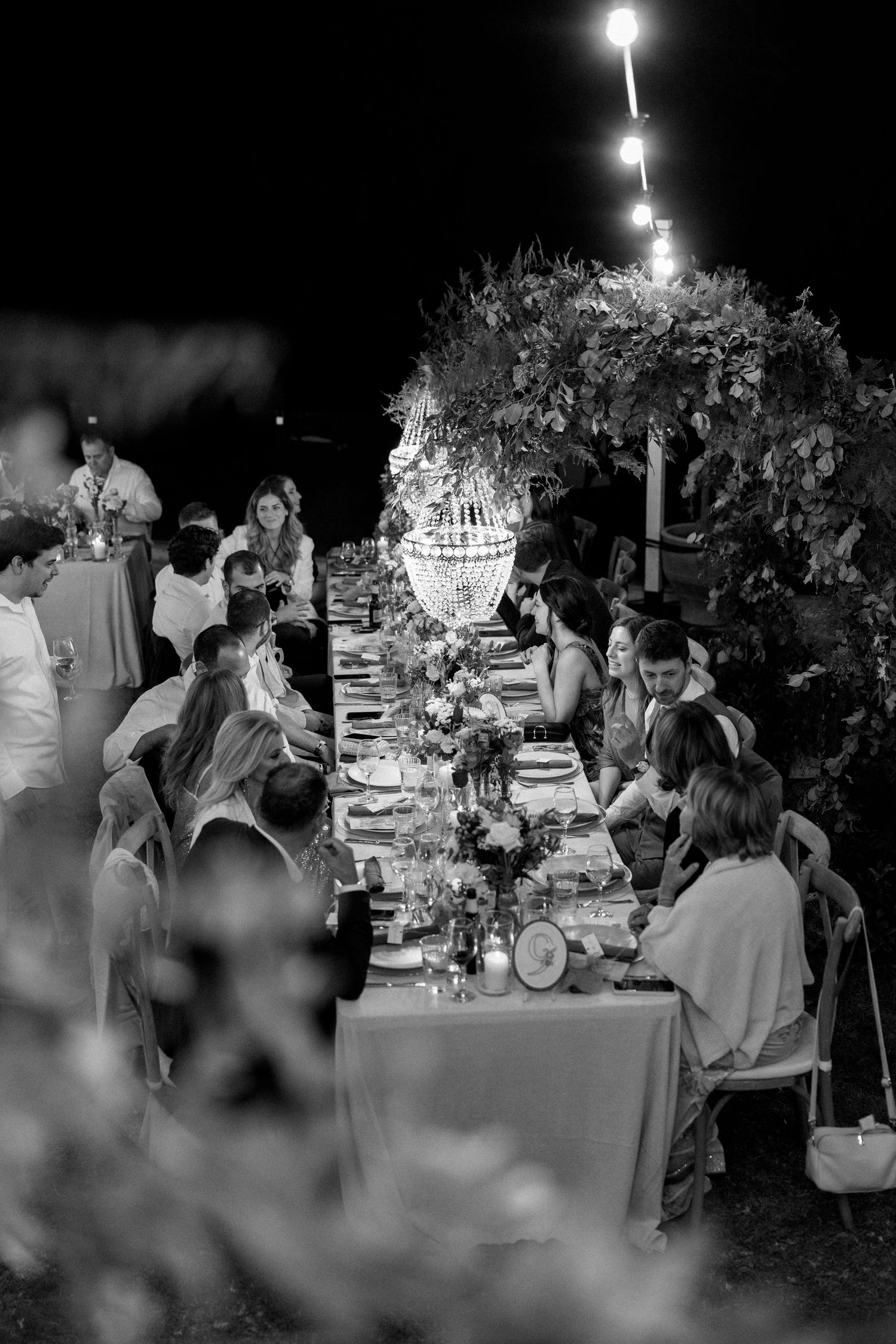Elegantly decorated outdoor wedding reception dinner table under string lights with floral arrangements and candlelight.