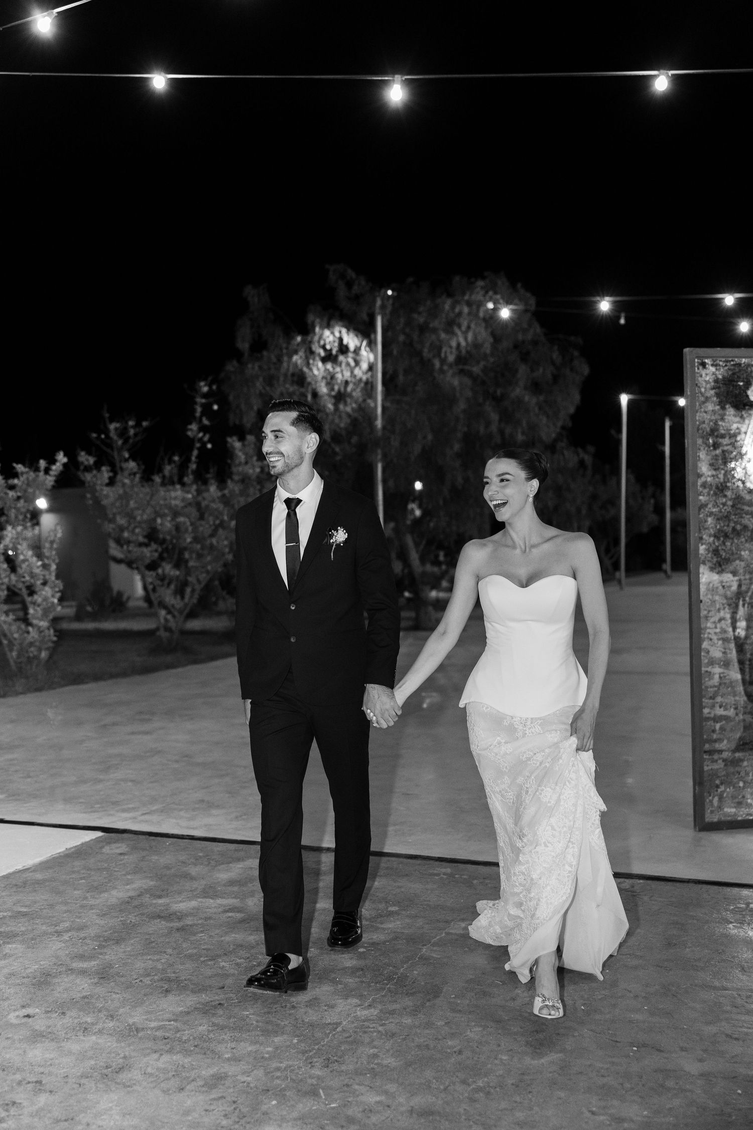 Black and white photo of newlyweds walking hand in hand under string lights at outdoor reception.