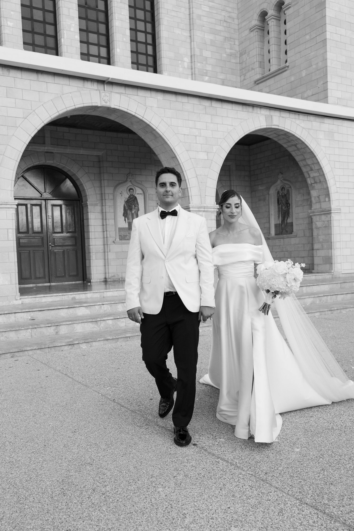 Black and white photo of a bride and groom walking hand in hand outside a stone church after their wedding ceremony.