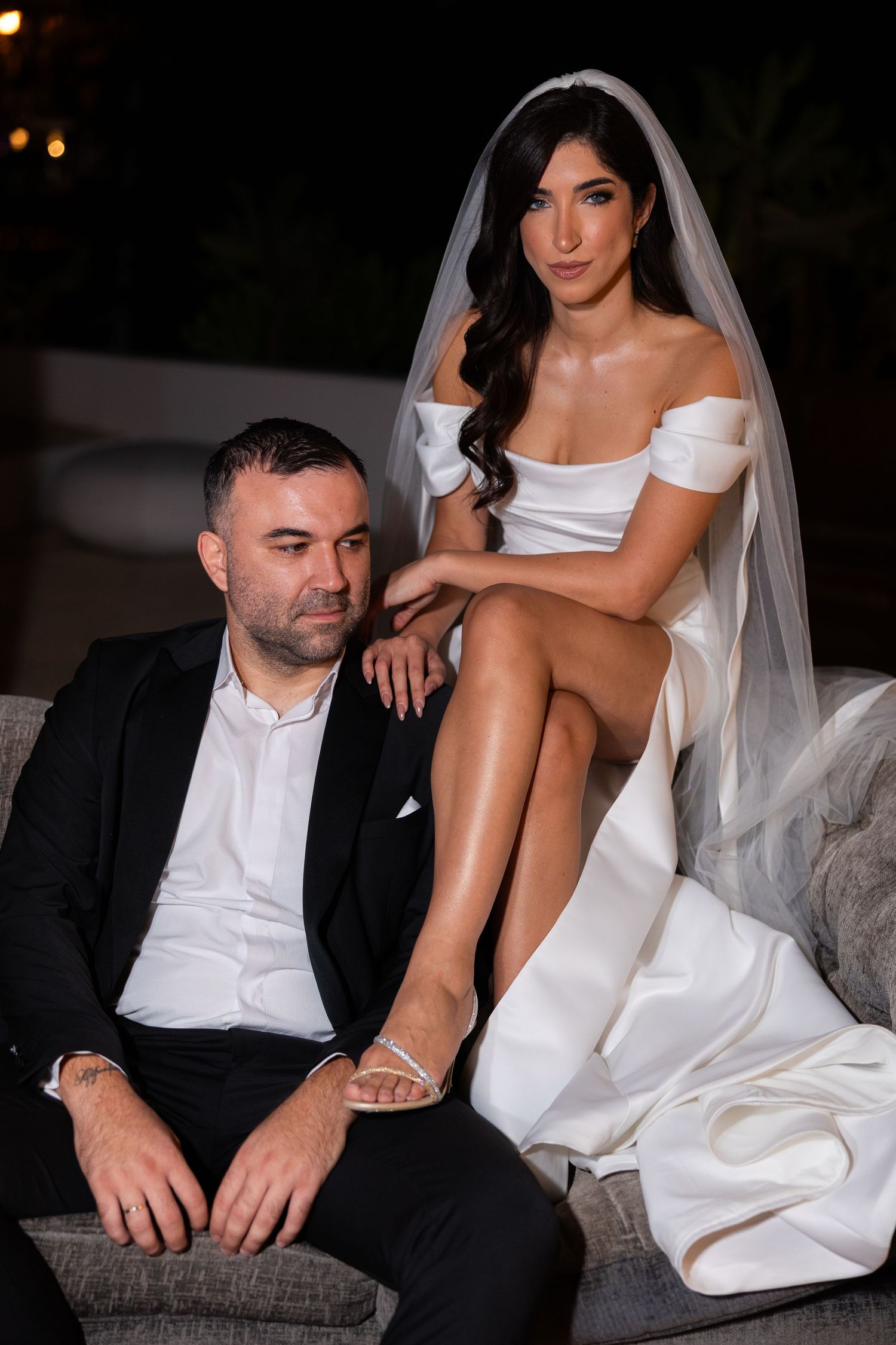Bride in white off-shoulder gown with veil poses beside groom in black suit on outdoor sofa at night.