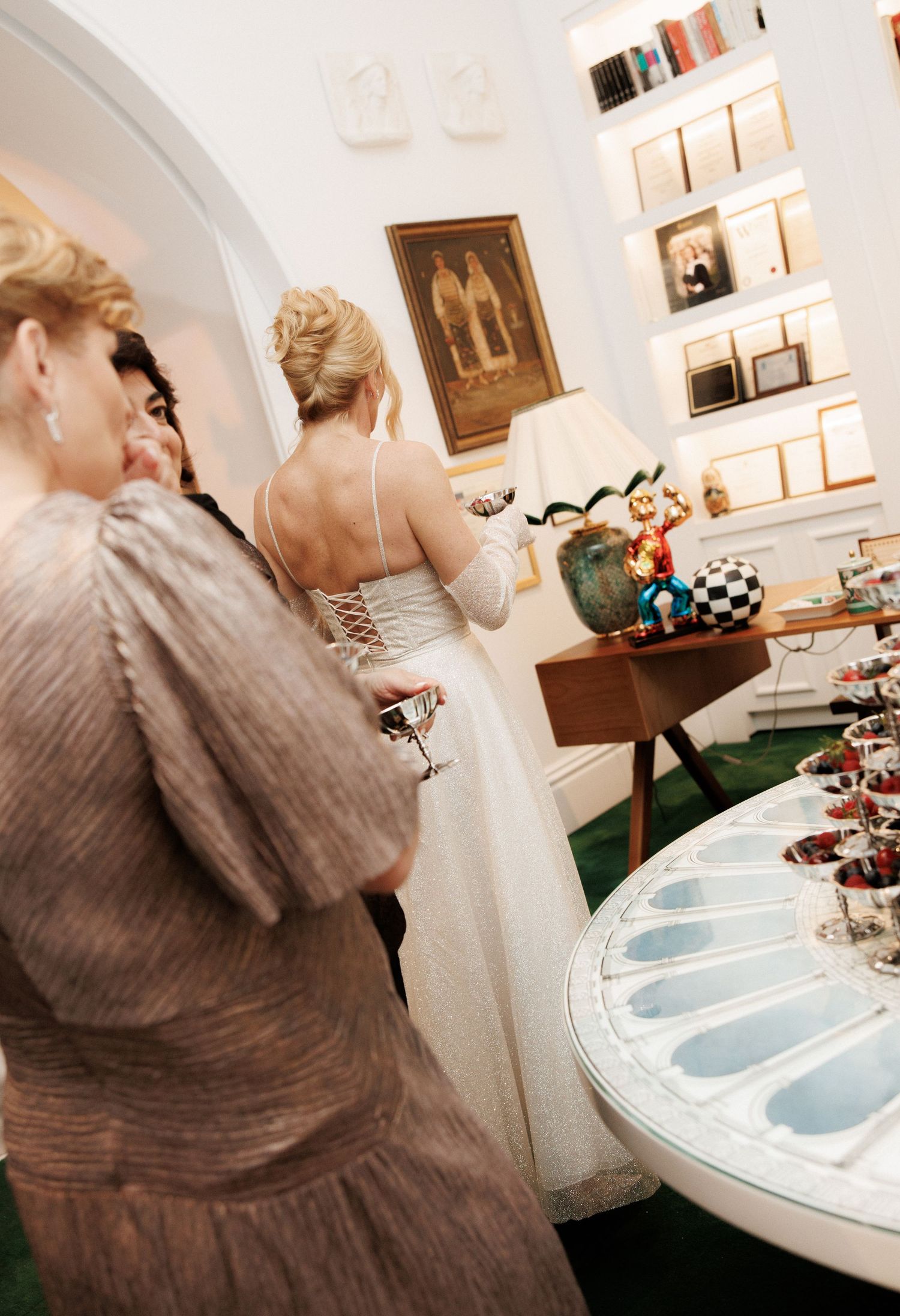 Bride in white lace gown at elegant indoor reception with guests, decorative shelves, and dessert table.