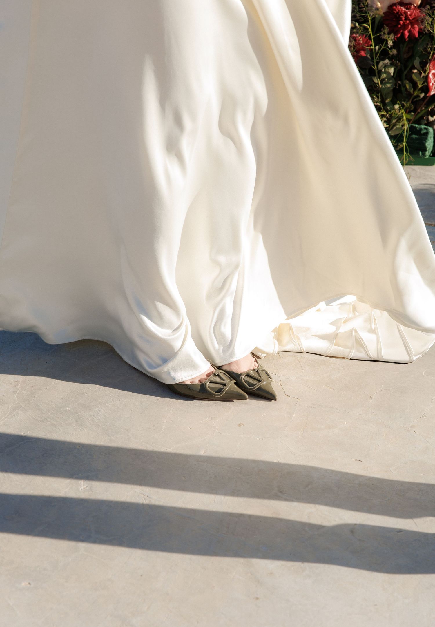 Close-up of a bride wearing an ivory satin wedding gown and olive green pointed-toe flats on a concrete surface.