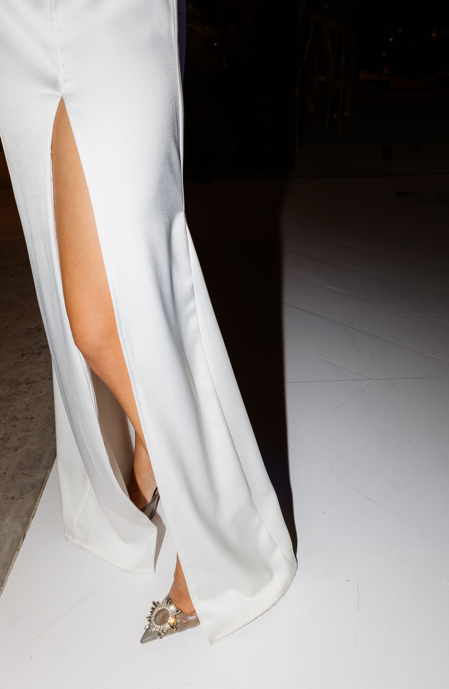 White gown with high front slit and embellished pointed-toe heels on light floor.