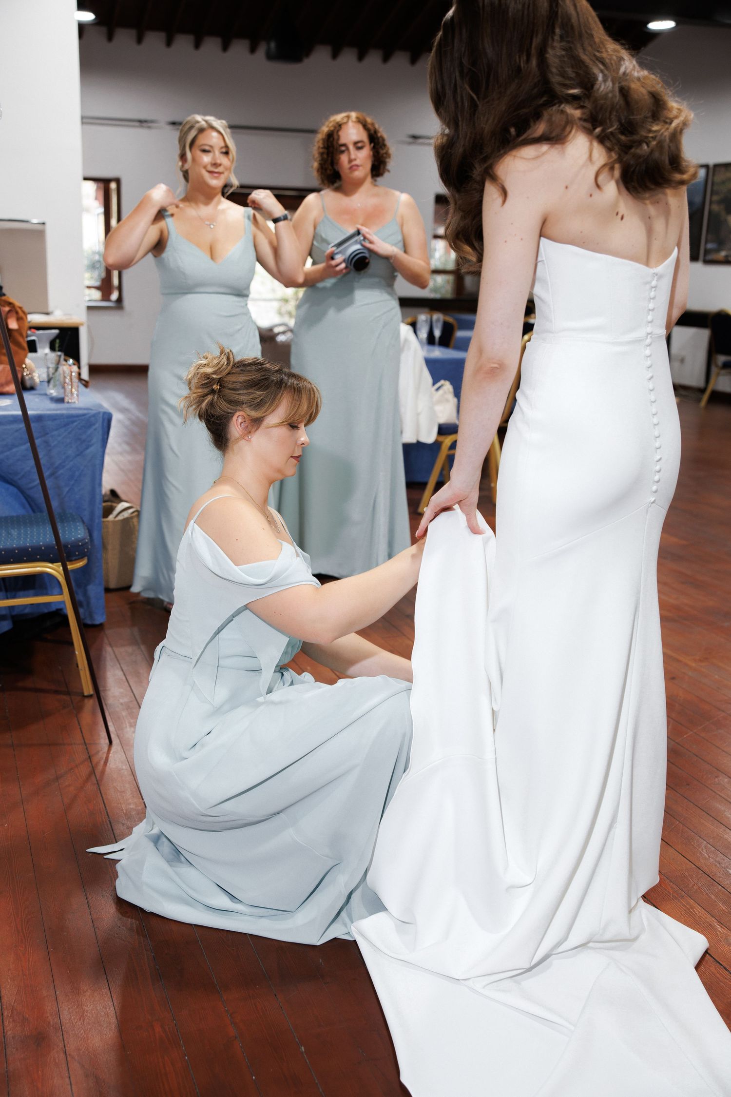 Bridesmaids in light blue dresses help adjust a white wedding gown during pre-ceremony preparations.