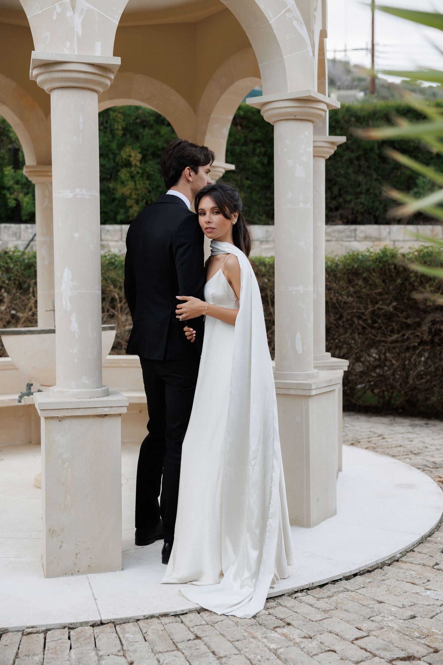 A bride and groom embrace beneath elegant stone arches during their romantic wedding ceremony.