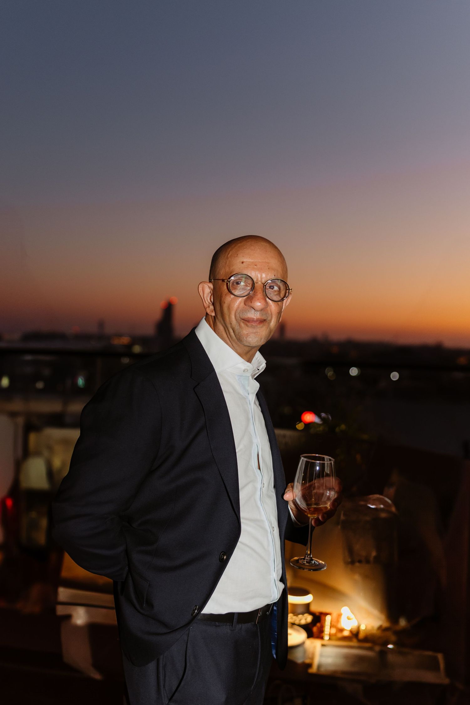 Person in formal attire holding wine glass on rooftop terrace during sunset.
