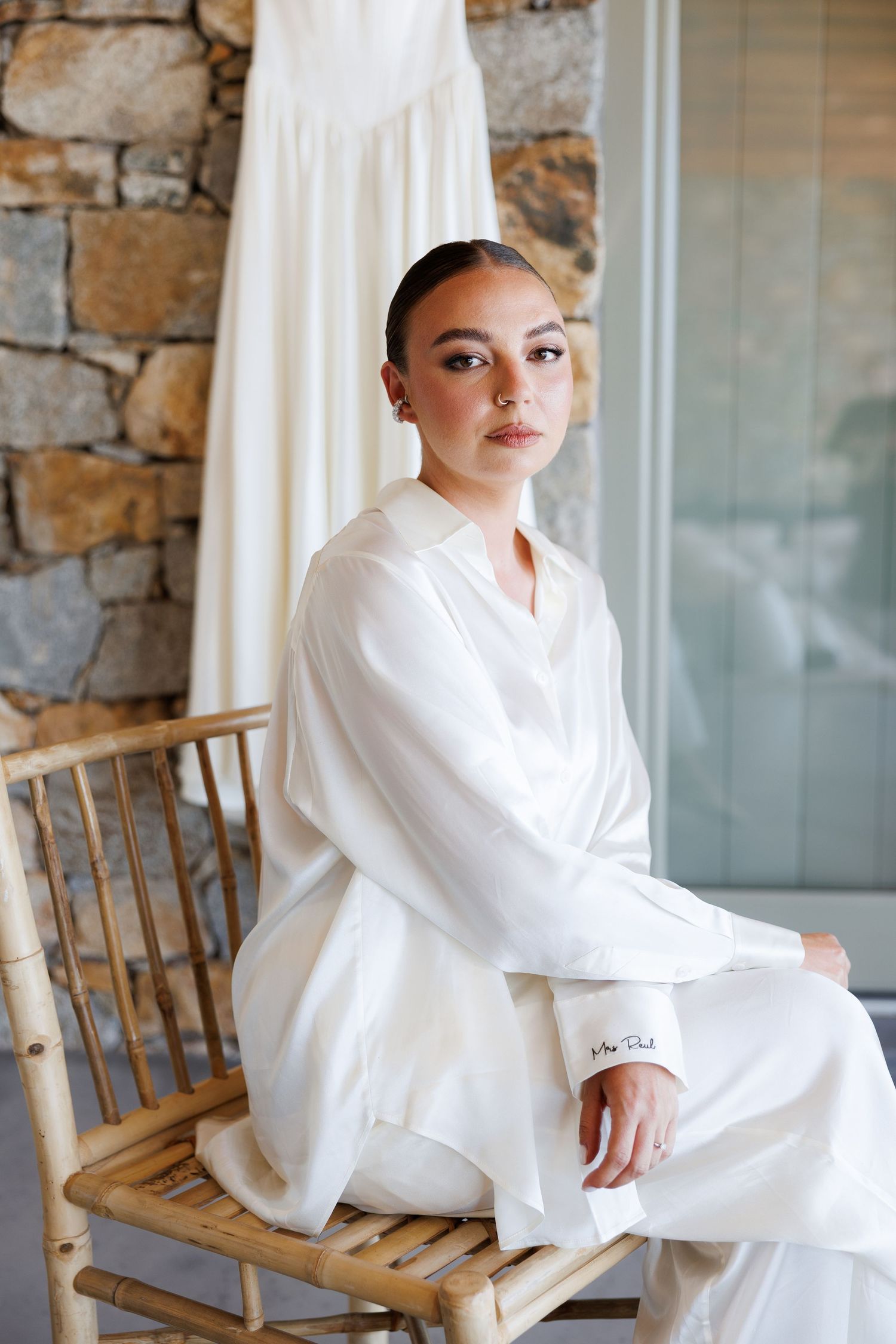 Bride in white silk robe sits on bamboo chair before wedding ceremony in front of stone wall.