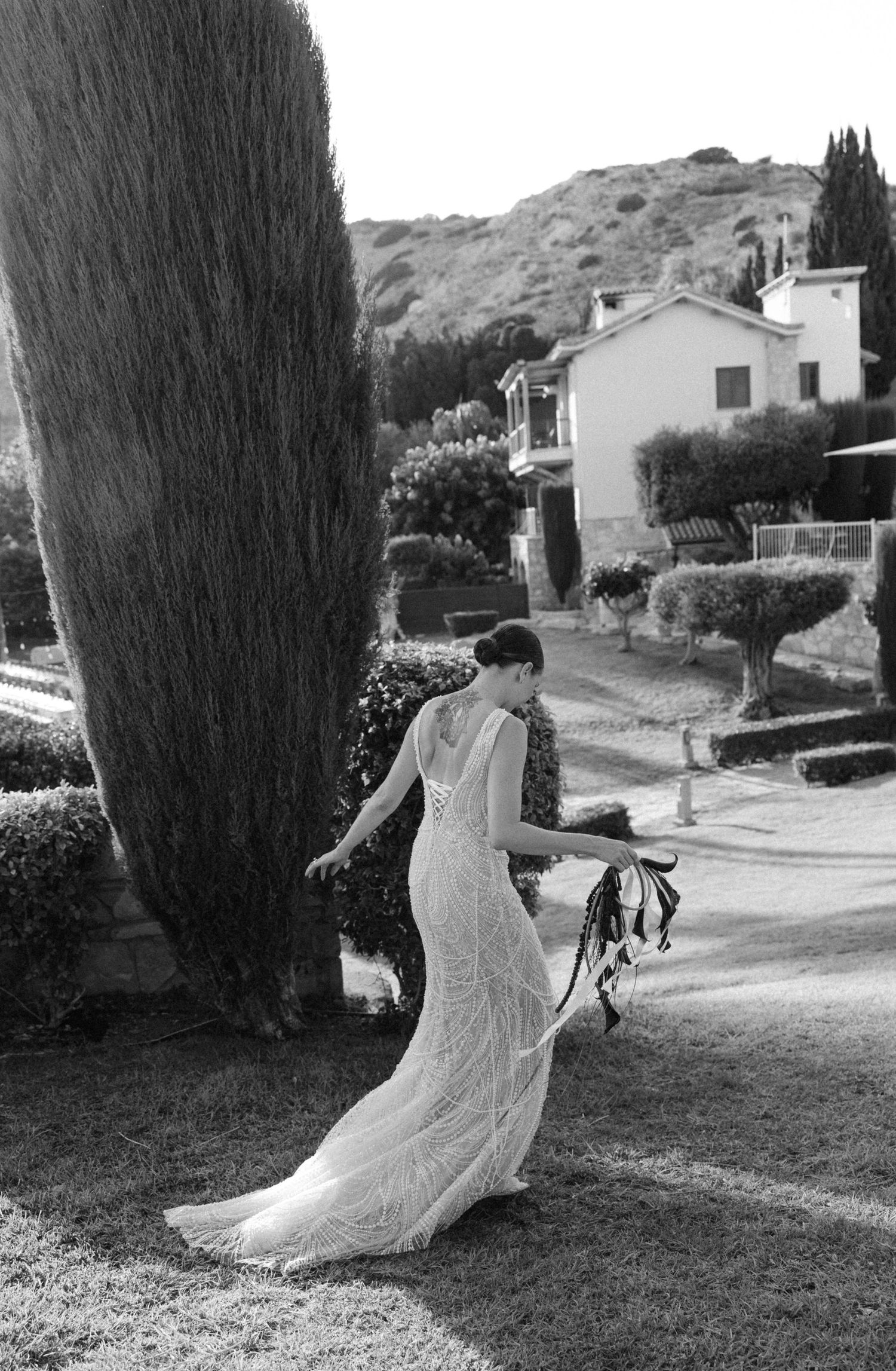 Bride in elegant lace wedding gown standing by tall cypress tree in scenic outdoor garden setting.