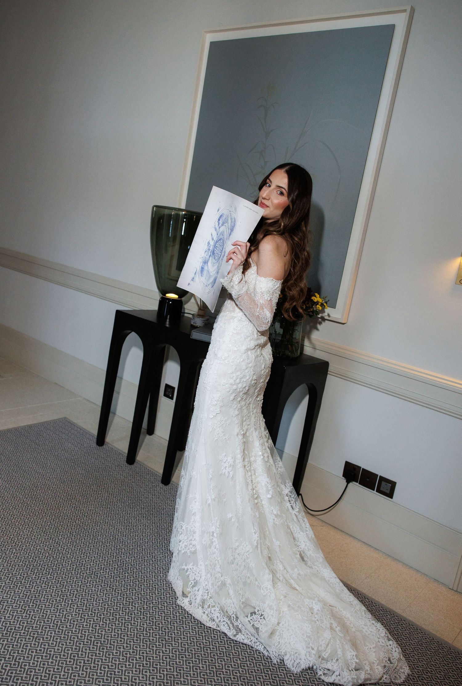 Bride in lace mermaid wedding gown holding floral artwork in elegant room with dark console table.