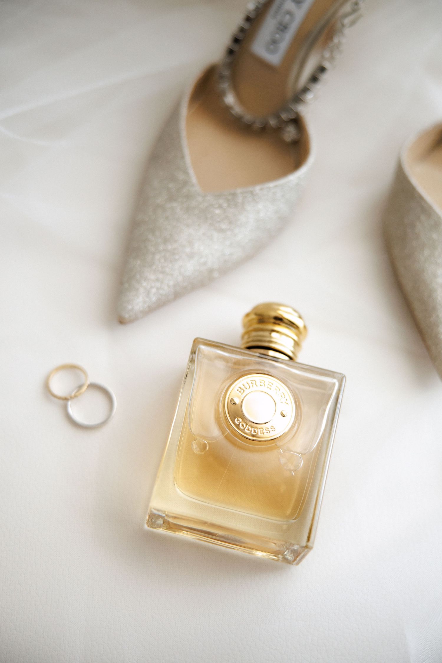 Gold perfume bottle with wedding rings and glittery Jimmy Choo bridal heels on white fabric background.