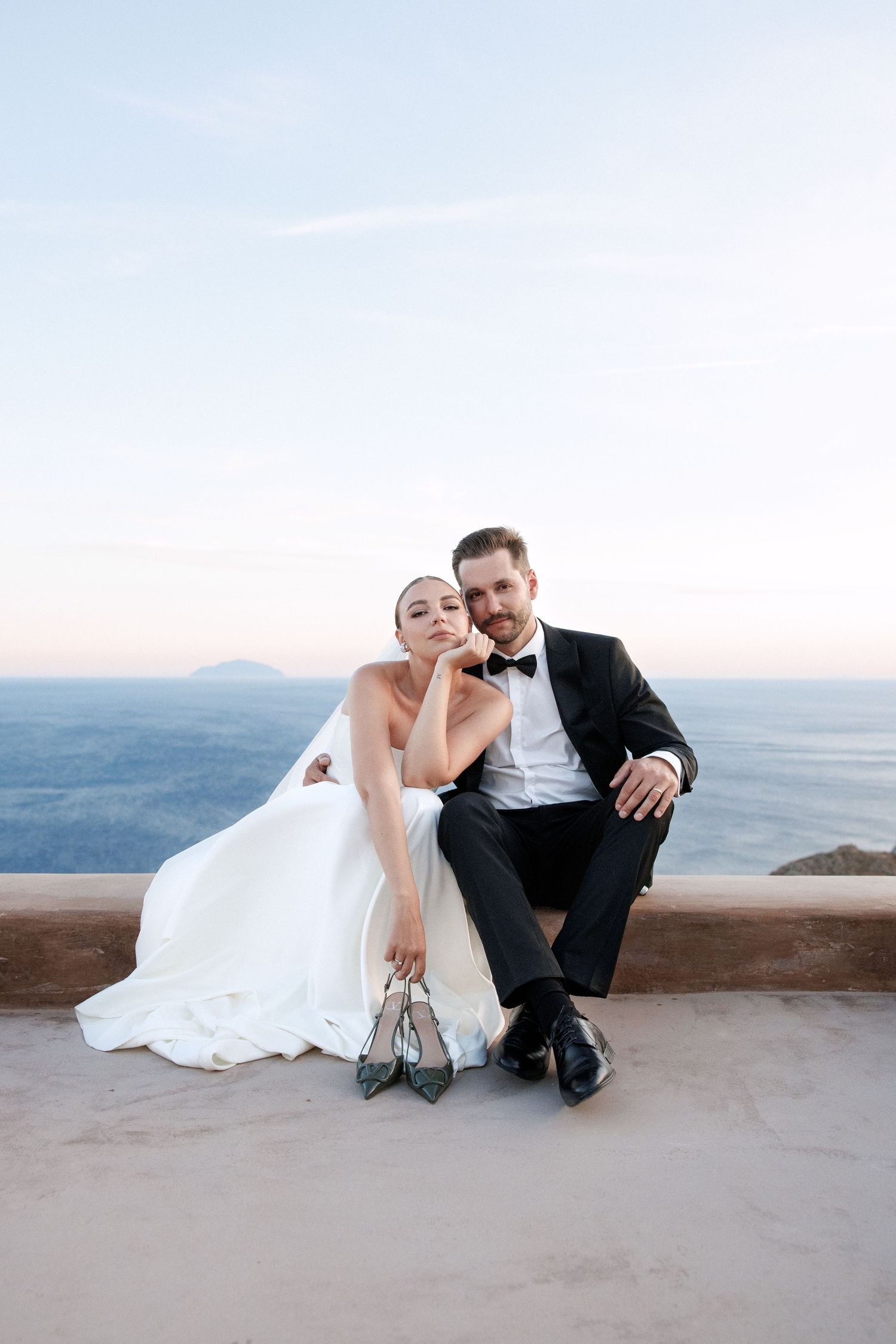 Bride and groom sitting together on terrace with ocean view, bride holding heels at sunset.
