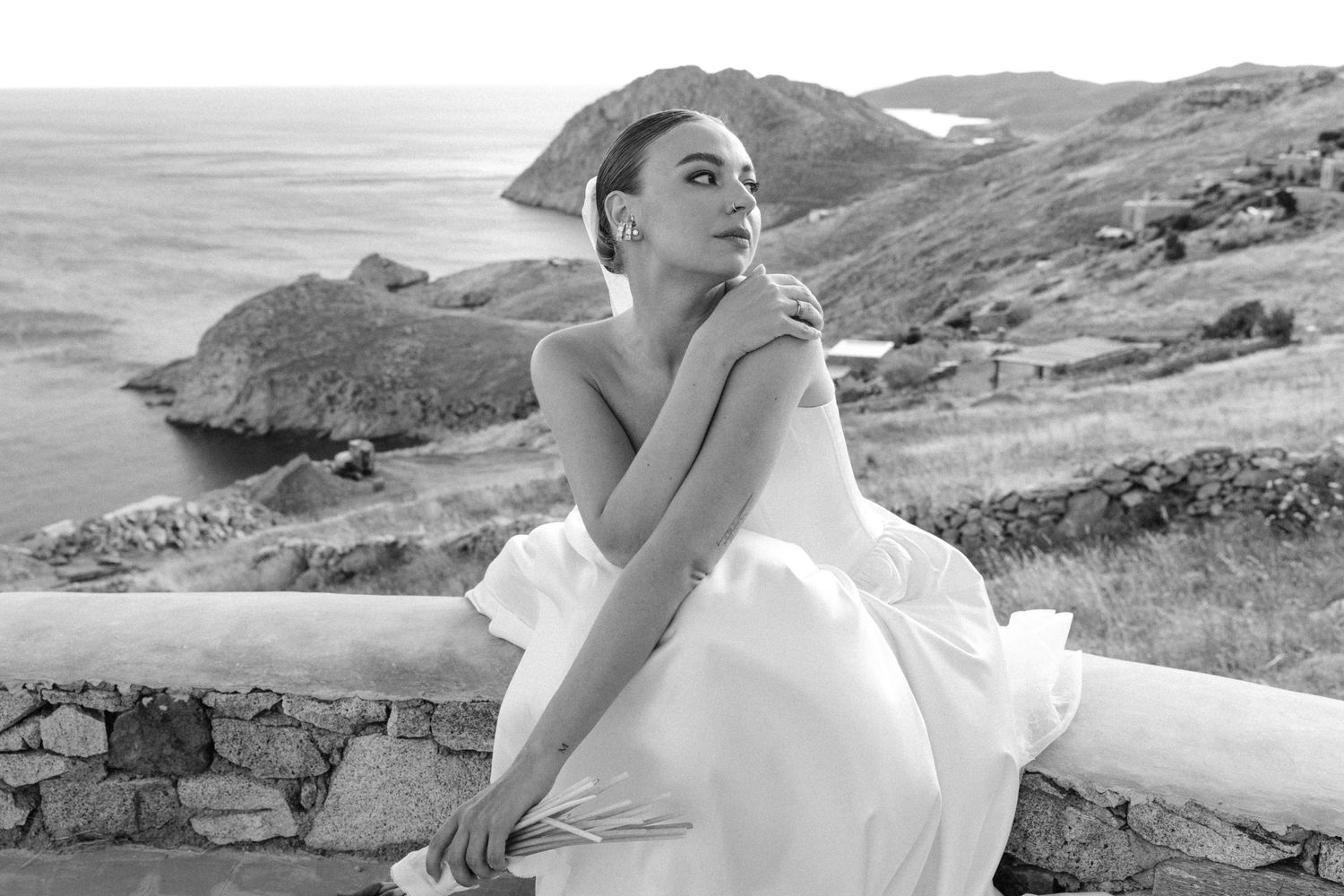 Elegant bride in a white gown poses gracefully on a stone wall with a stunning coastal hillside backdrop.
