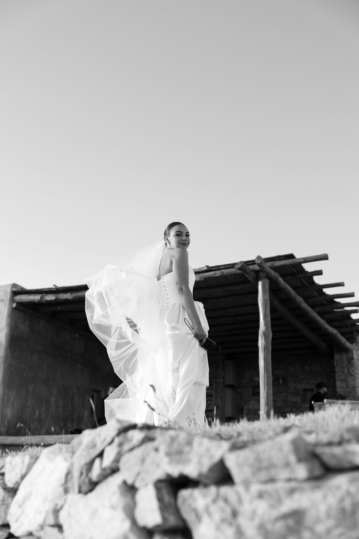 Black and white photo of a bride in flowing wedding dress and veil standing on rocks near rustic wooden structure.
