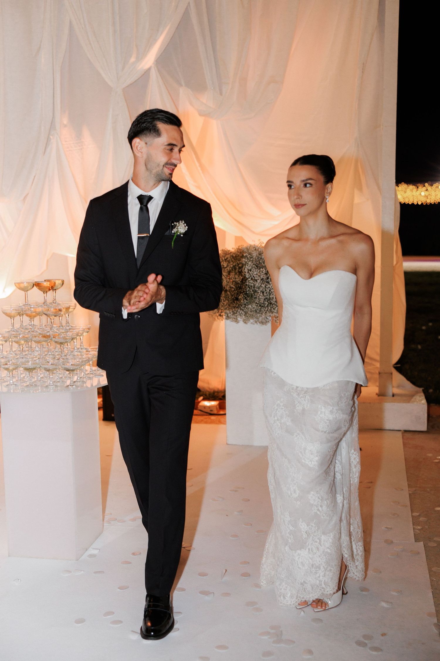 A wedding couple walking together in formal attire against a draped white backdrop.