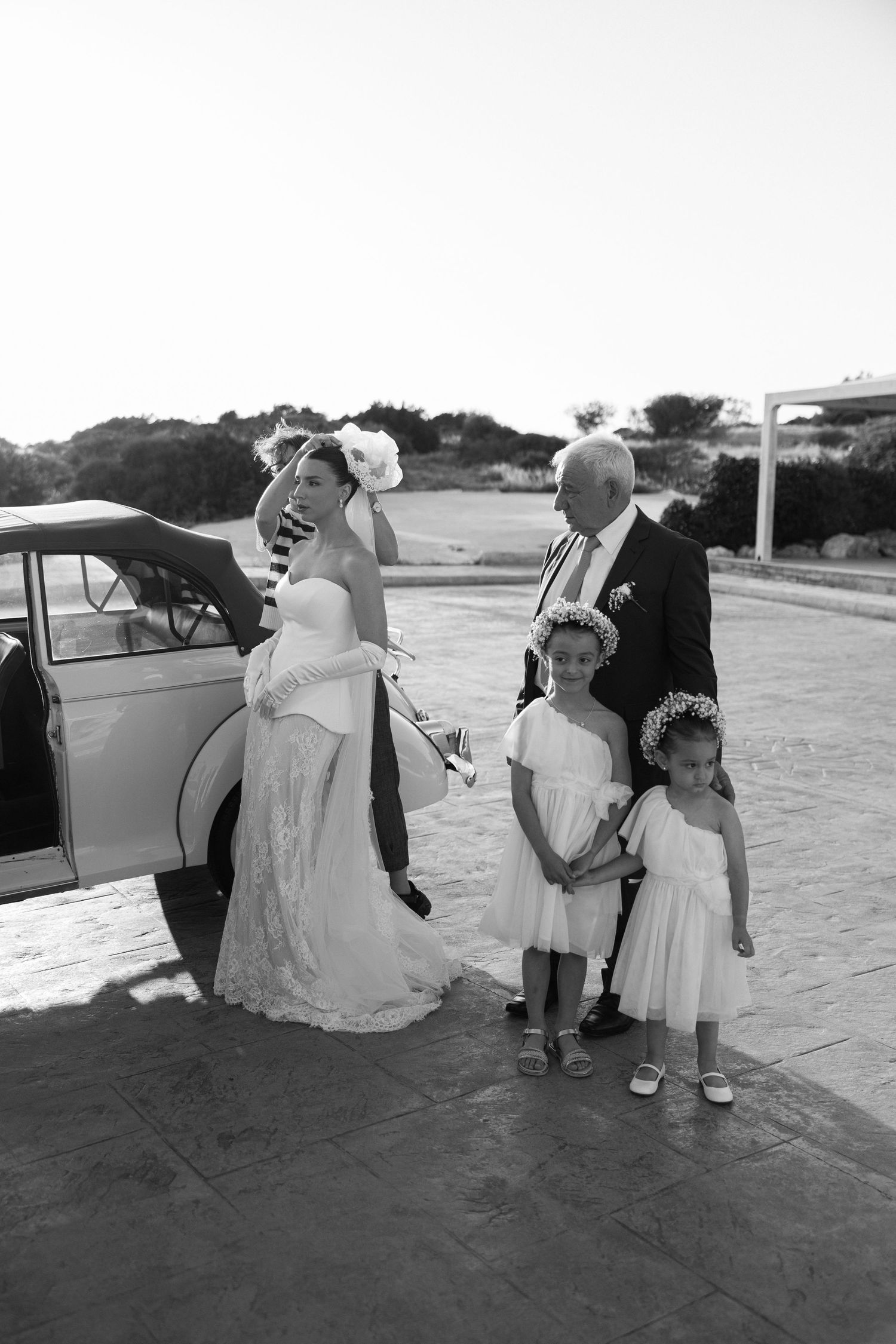 Black and white wedding photo with a bride and family near a vintage car.