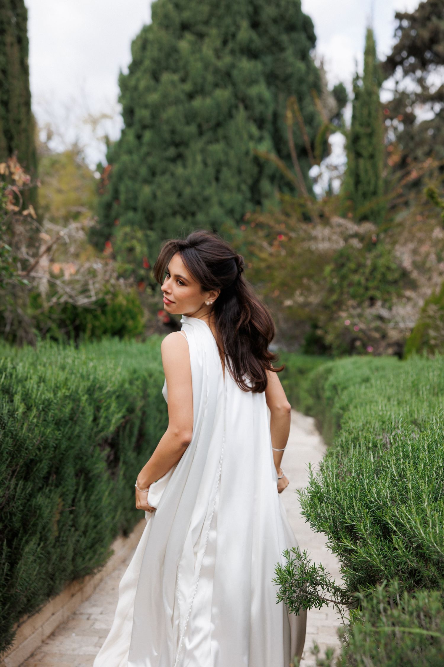 Woman in flowing white dress poses on garden path surrounded by rosemary and cypress trees.