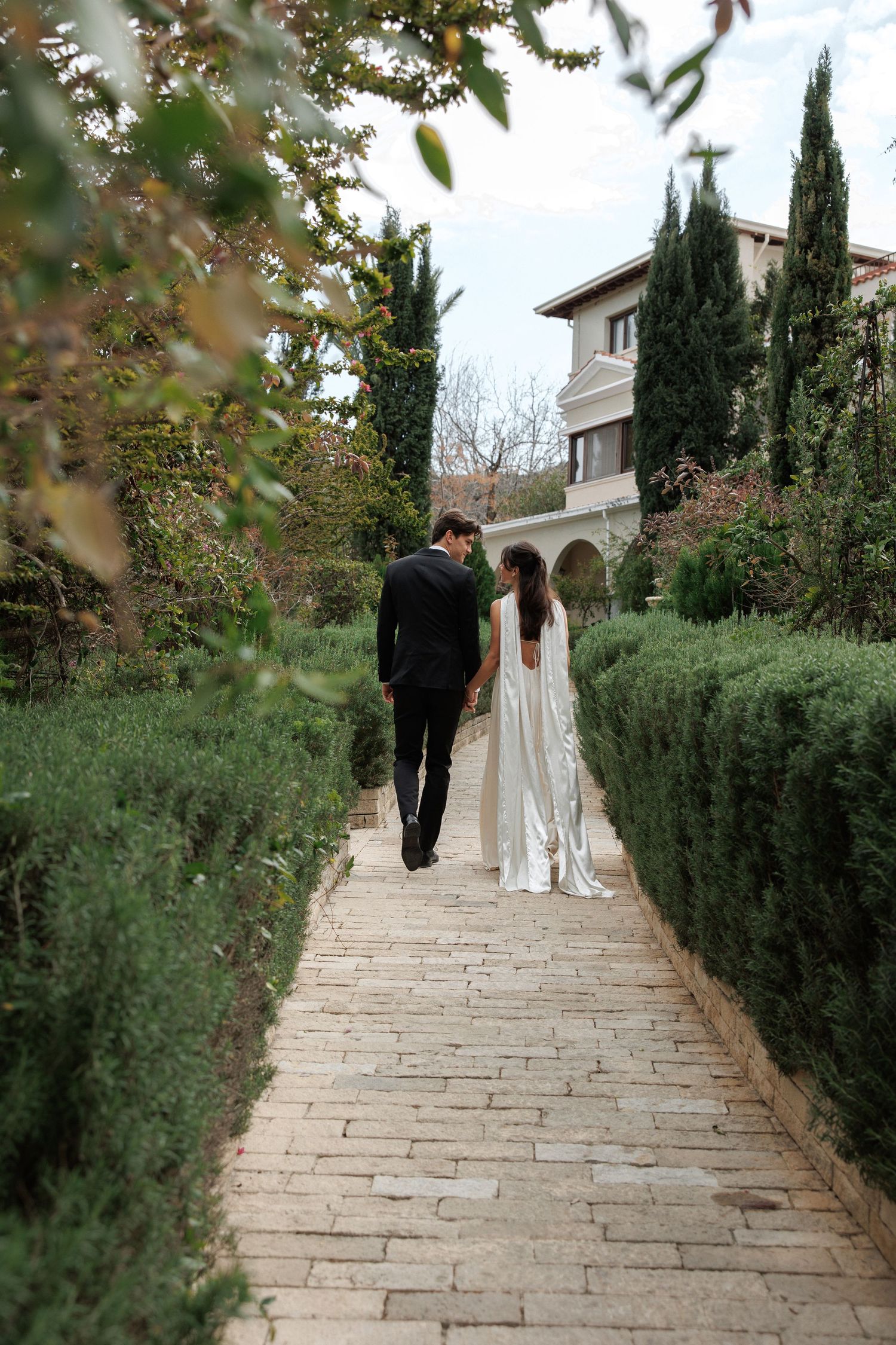 Newlyweds walking down a stone path lined with cypress trees and greenery at an elegant venue.