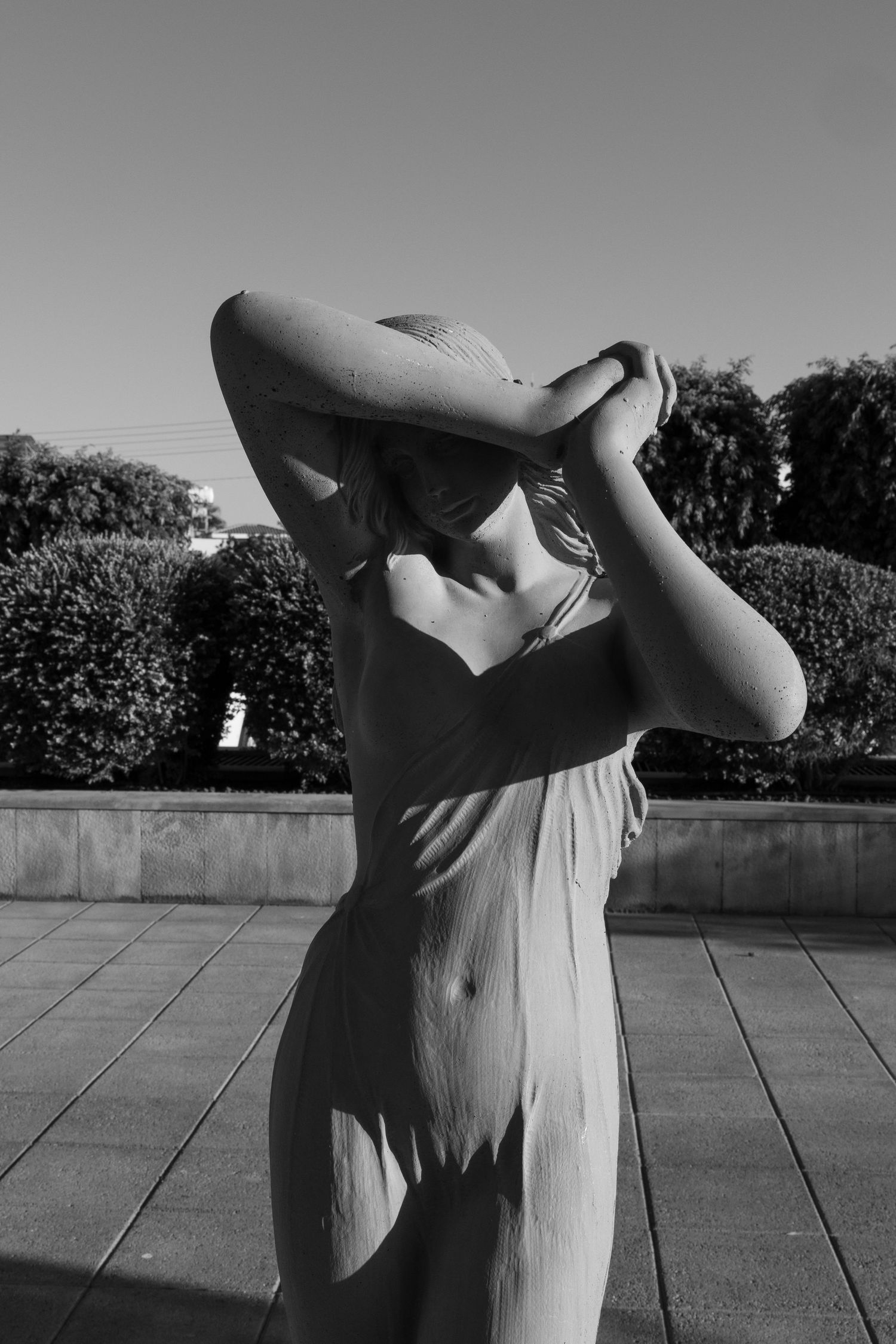 Black and white photo of a classical female sculpture outdoors, arms raised, with hedges and clear sky background.