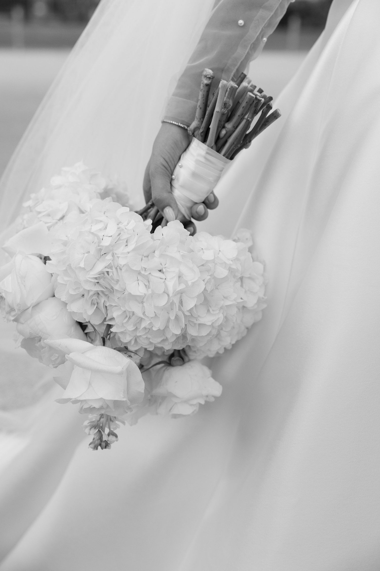 Black and white photo of a bride holding a white hydrangea wedding bouquet with flowing veil in background.
