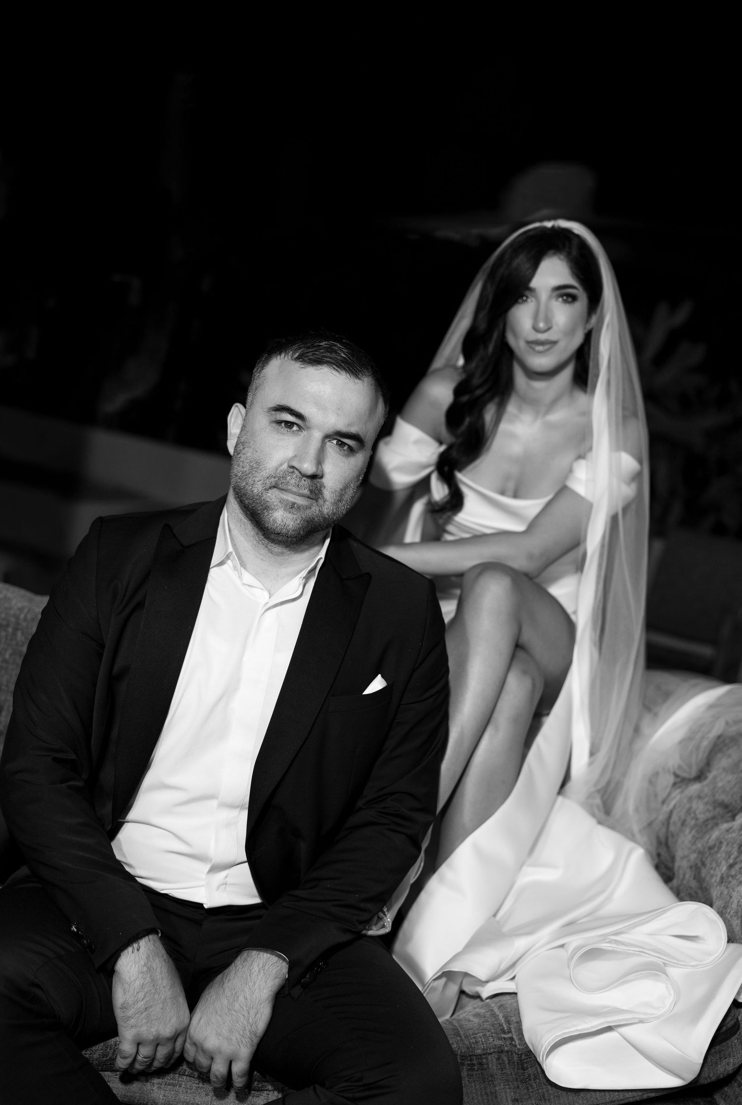 Black and white wedding portrait of a groom in a suit seated with his bride in a veil and gown behind him.