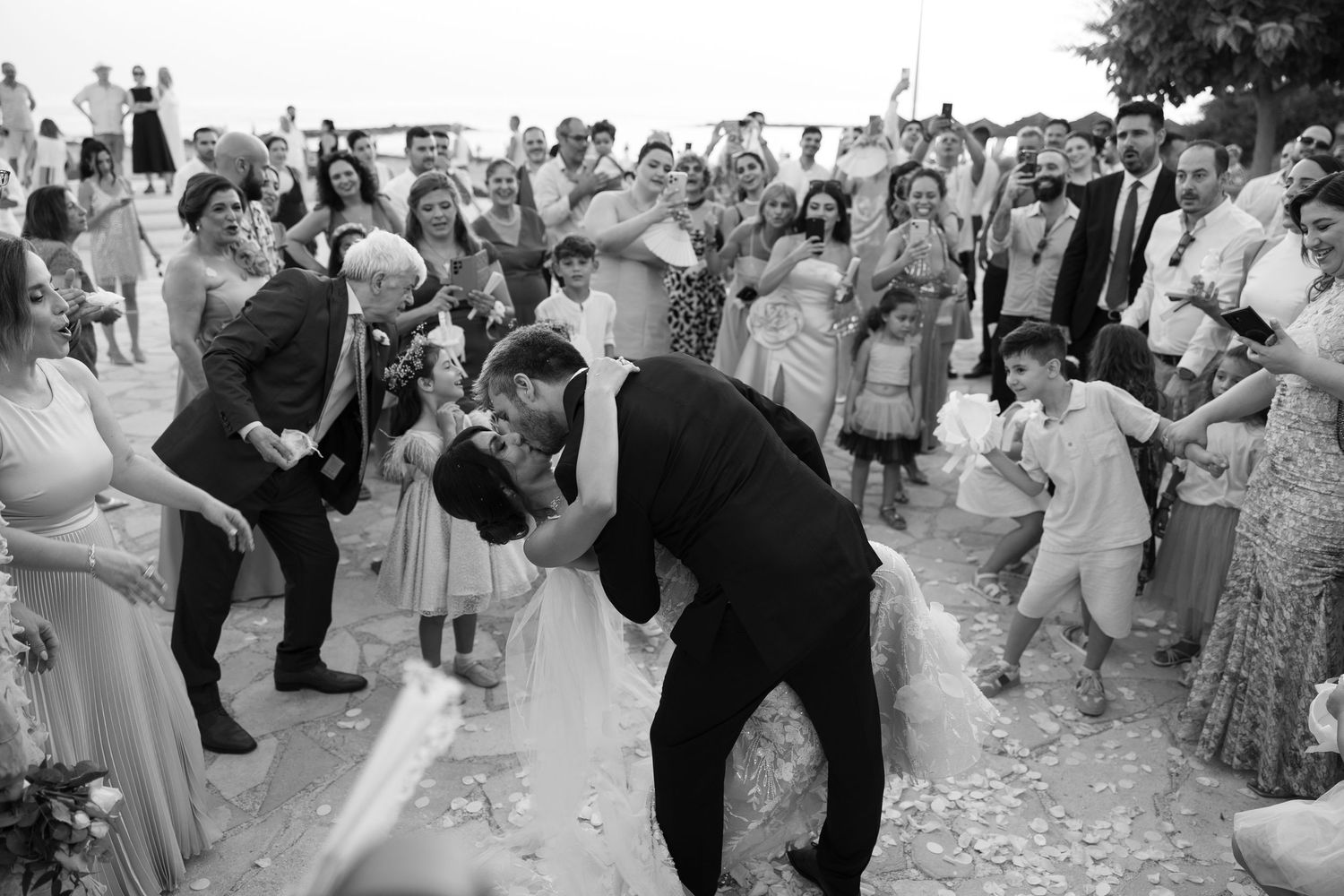 Black and white photo of bride and groom sharing a kiss surrounded by cheering wedding guests outdoors.
