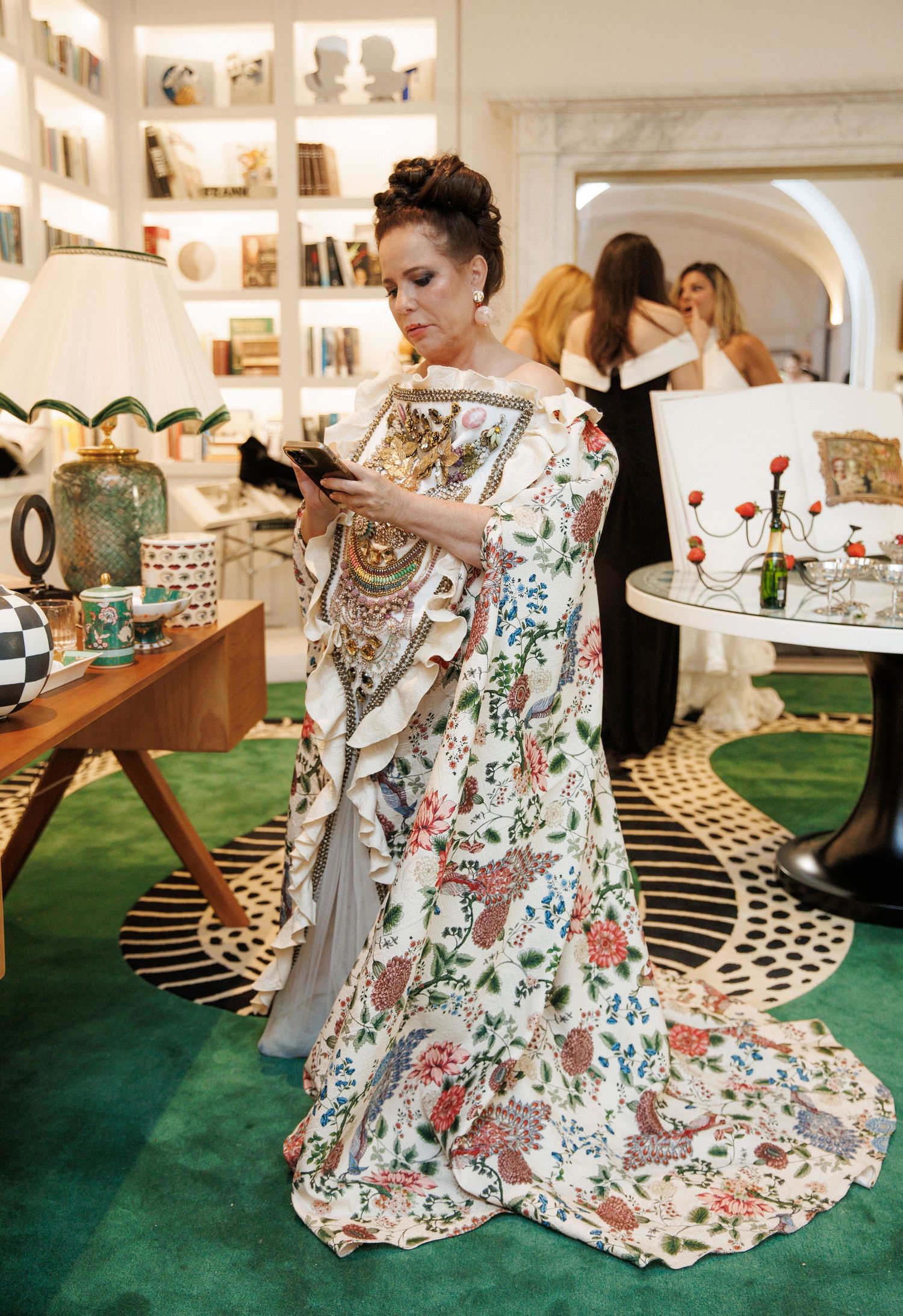 Woman in elegant floral off-shoulder gown checks phone at upscale boutique event with green carpet.