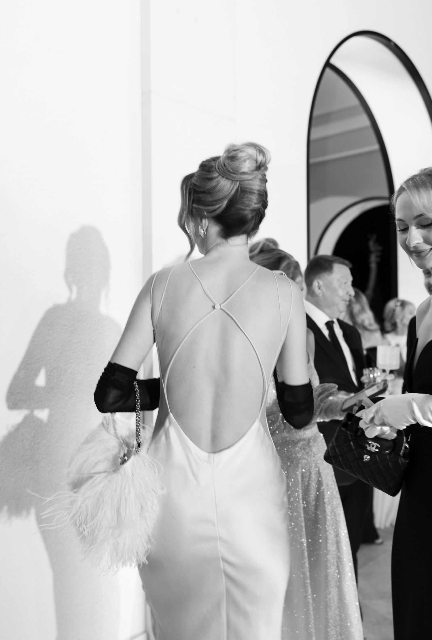 Black and white photo of woman in elegant backless gown with cross-strap detail, updo hairstyle, black gloves, feather clutch.