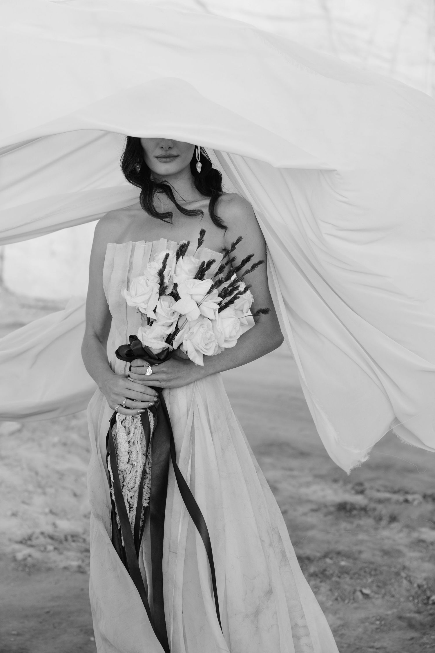 Black and white photo of a bride holding a white flower bouquet with fabric veil blowing over her face outdoors.