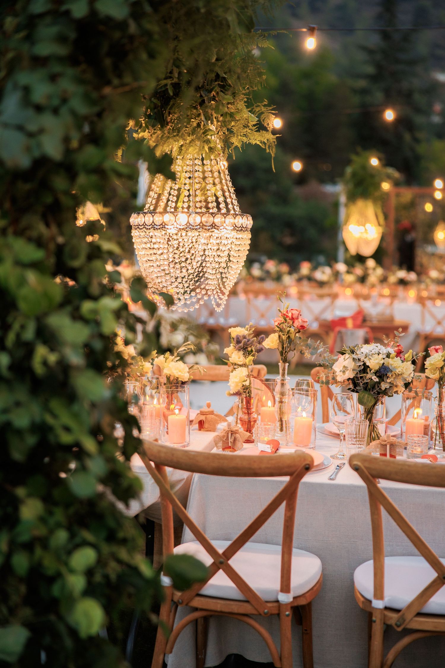 Crystal chandelier illuminates romantic outdoor table setting with floral centerpieces at dusk.