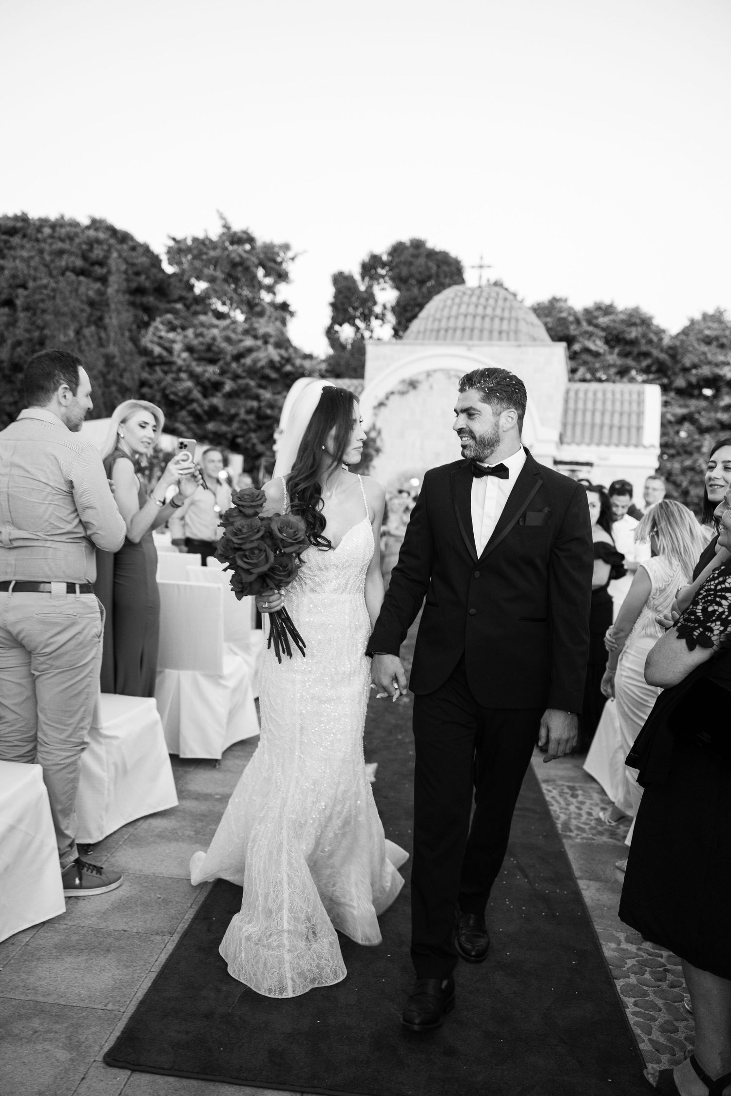 Black and white photo of bride and groom walking down the aisle at an outdoor wedding ceremony.