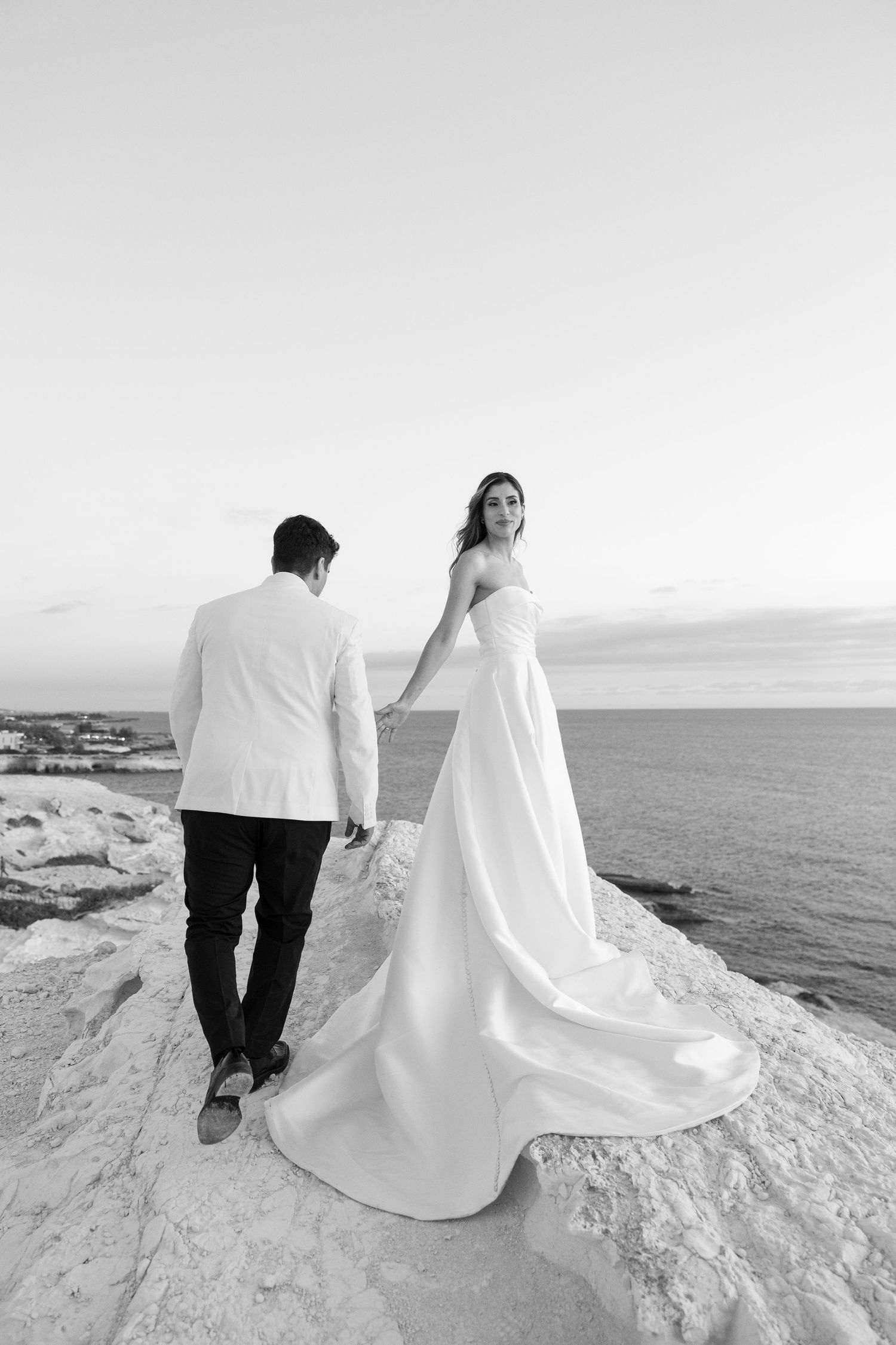 Black and white photo of bride and groom holding hands on rocky seaside cliff, bride's gown train flowing behind her.