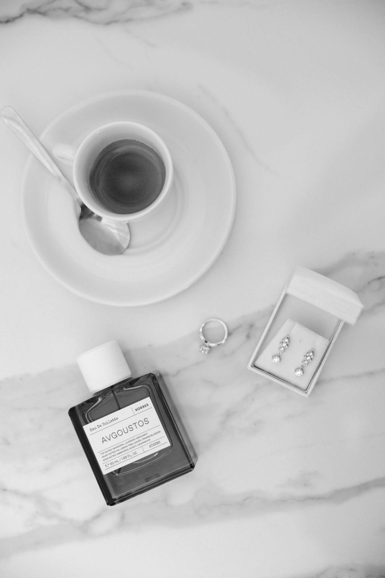 Black and white flat lay of coffee cup, Avgoustos perfume bottle, and jewelry box with earrings on marble.