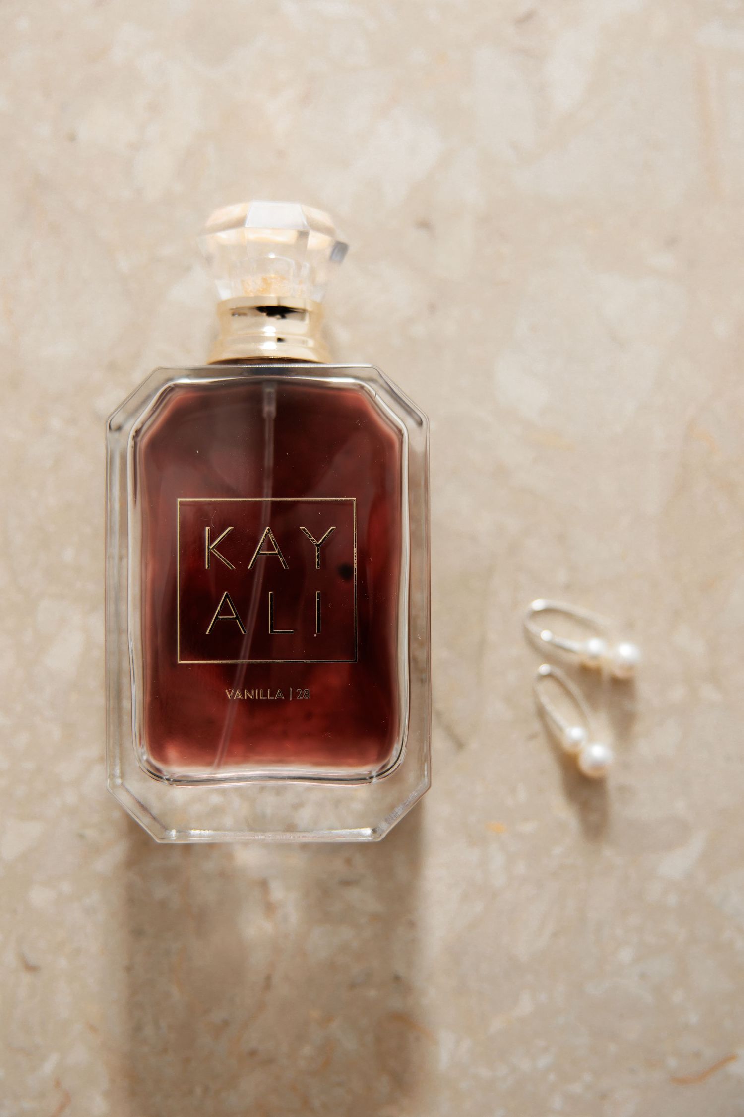 Kayali Vanilla 28 perfume bottle on marble surface beside pearl earrings.