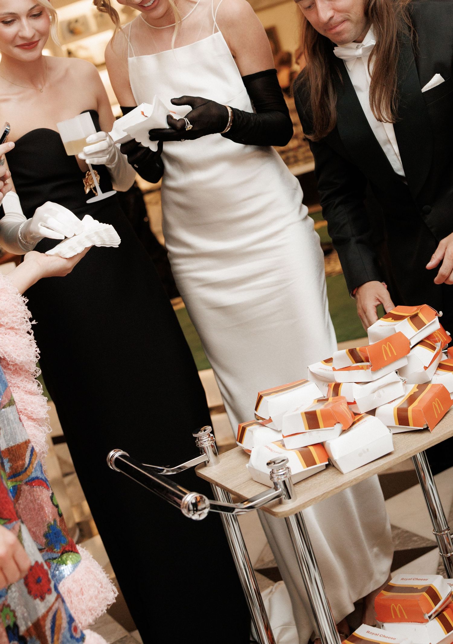 Elegant wedding guests in formal attire gather around a cart of McDonald's burgers at a chic reception.