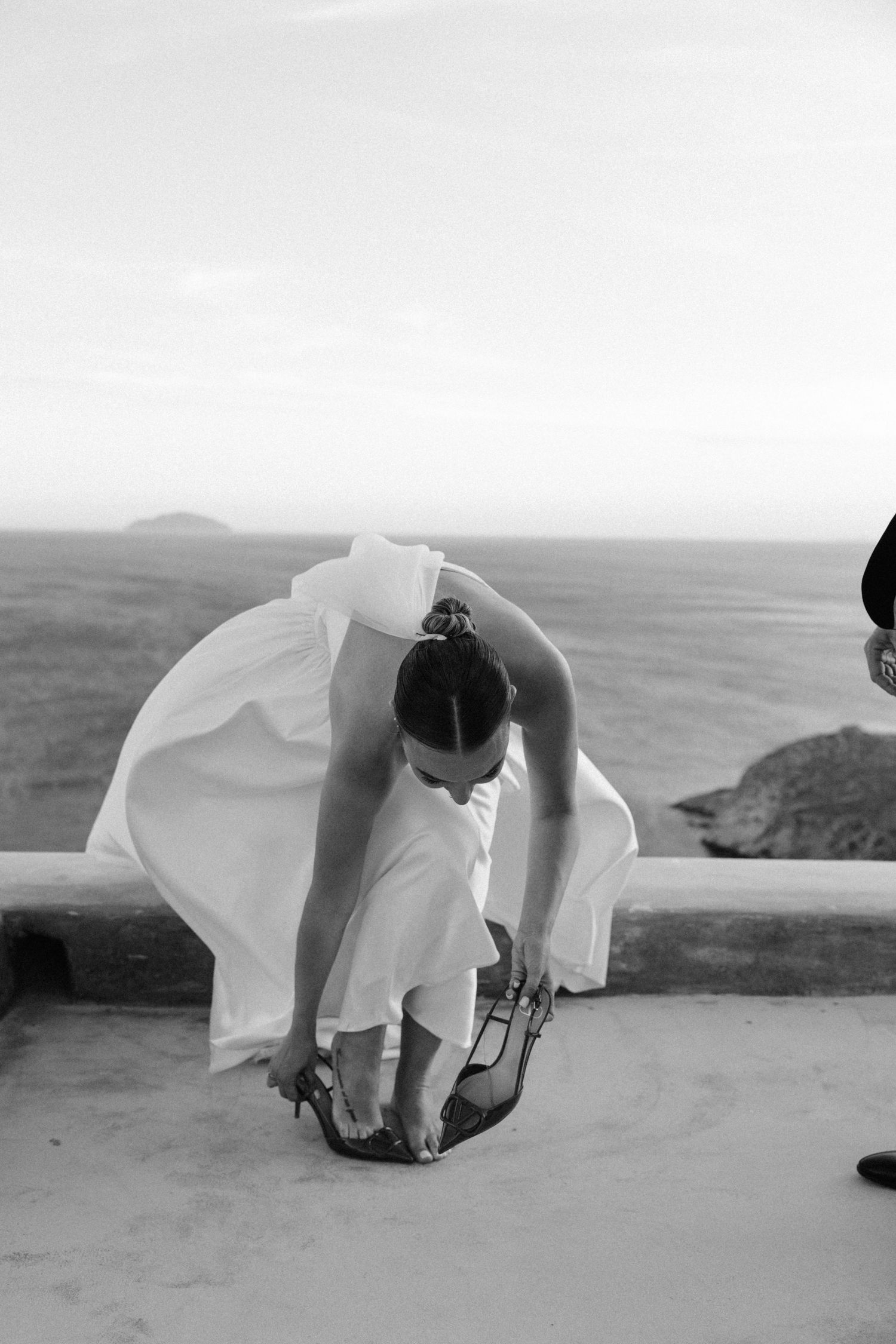 Bride bending to put on heels on seaside terrace in elegant black and white wedding photo.