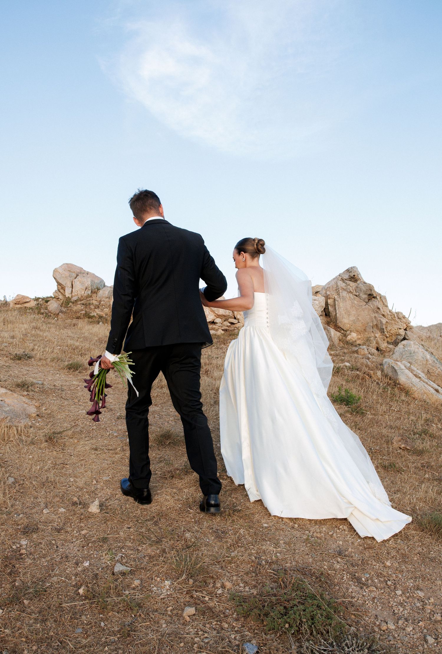 Bride and groom walking hand in hand on rocky hilltop, woman in white gown holding bouquet under clear blue sky.