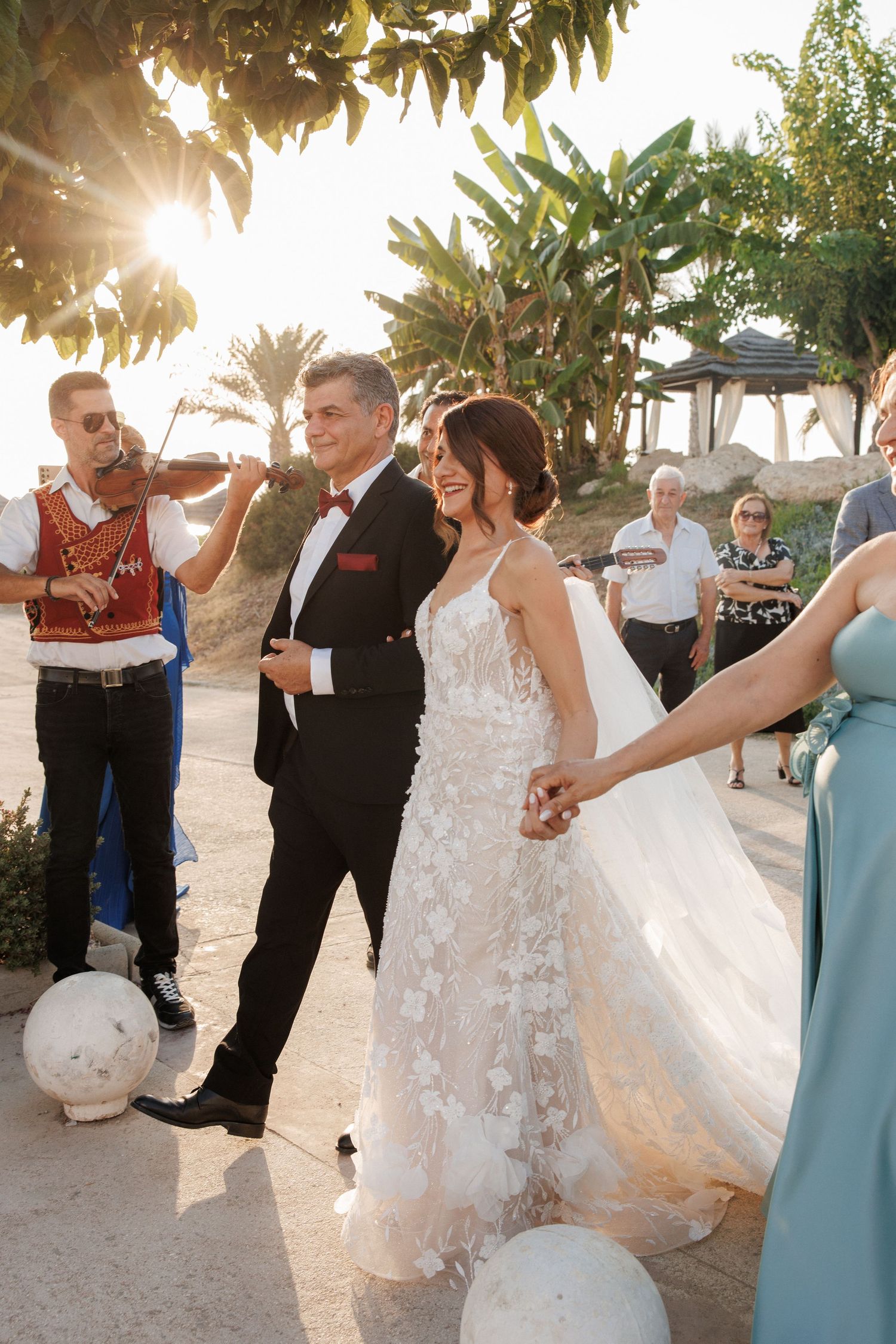 Bride and groom walk together outdoors at golden hour wedding, accompanied by violinist, with guests and tropical greenery in background.