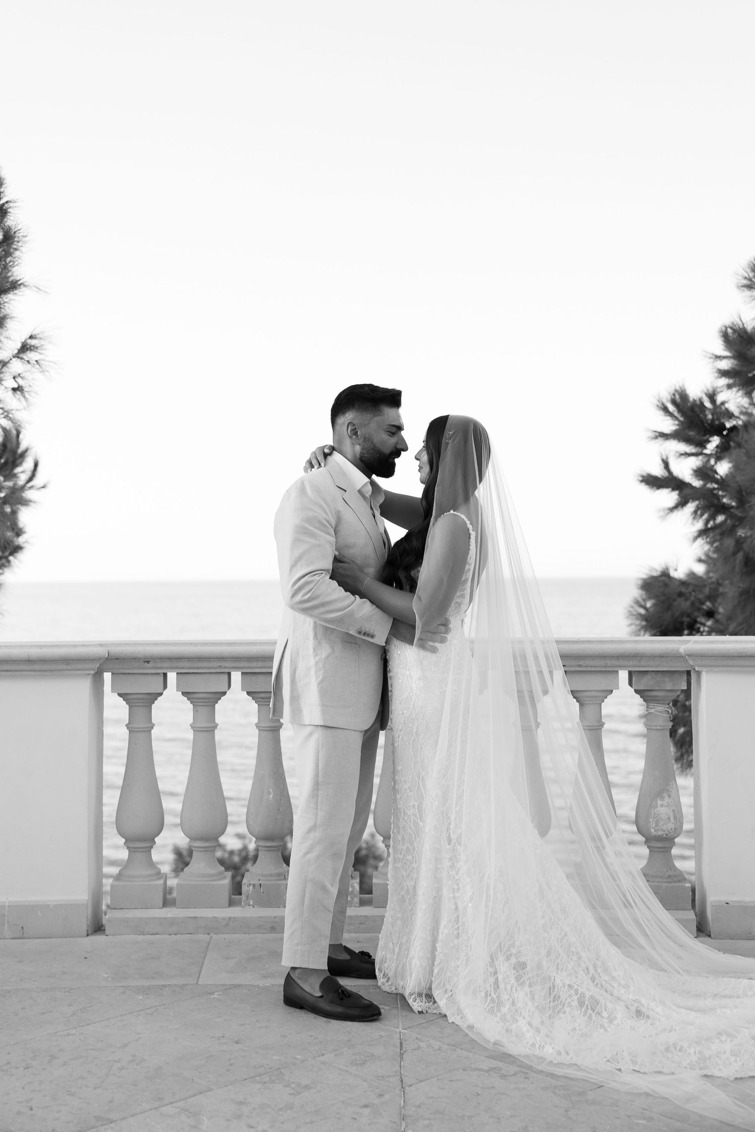 Black and white wedding portrait of bride in veil and groom embracing on balcony with ocean and palm trees behind them.