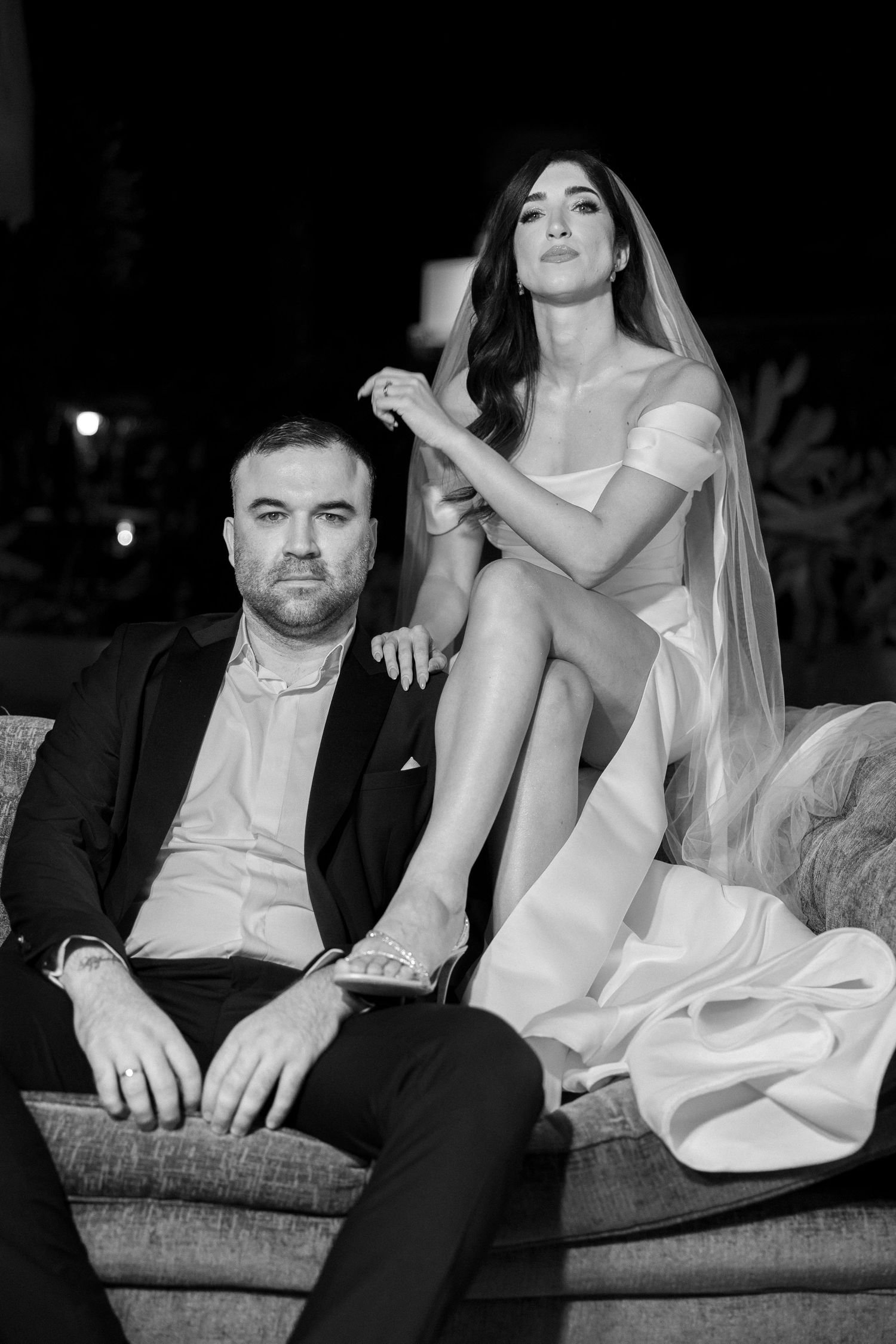 Black and white wedding photo of bride in veil and groom in suit sitting on sofa at night.