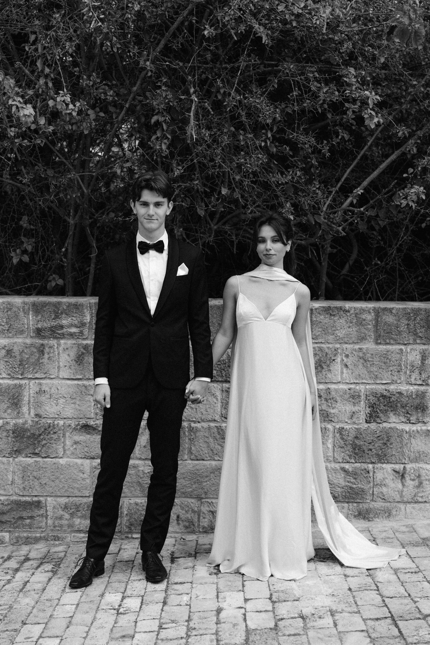 Black and white photo of a wedding couple standing together on brick pavers against a stone wall with foliage backdrop.