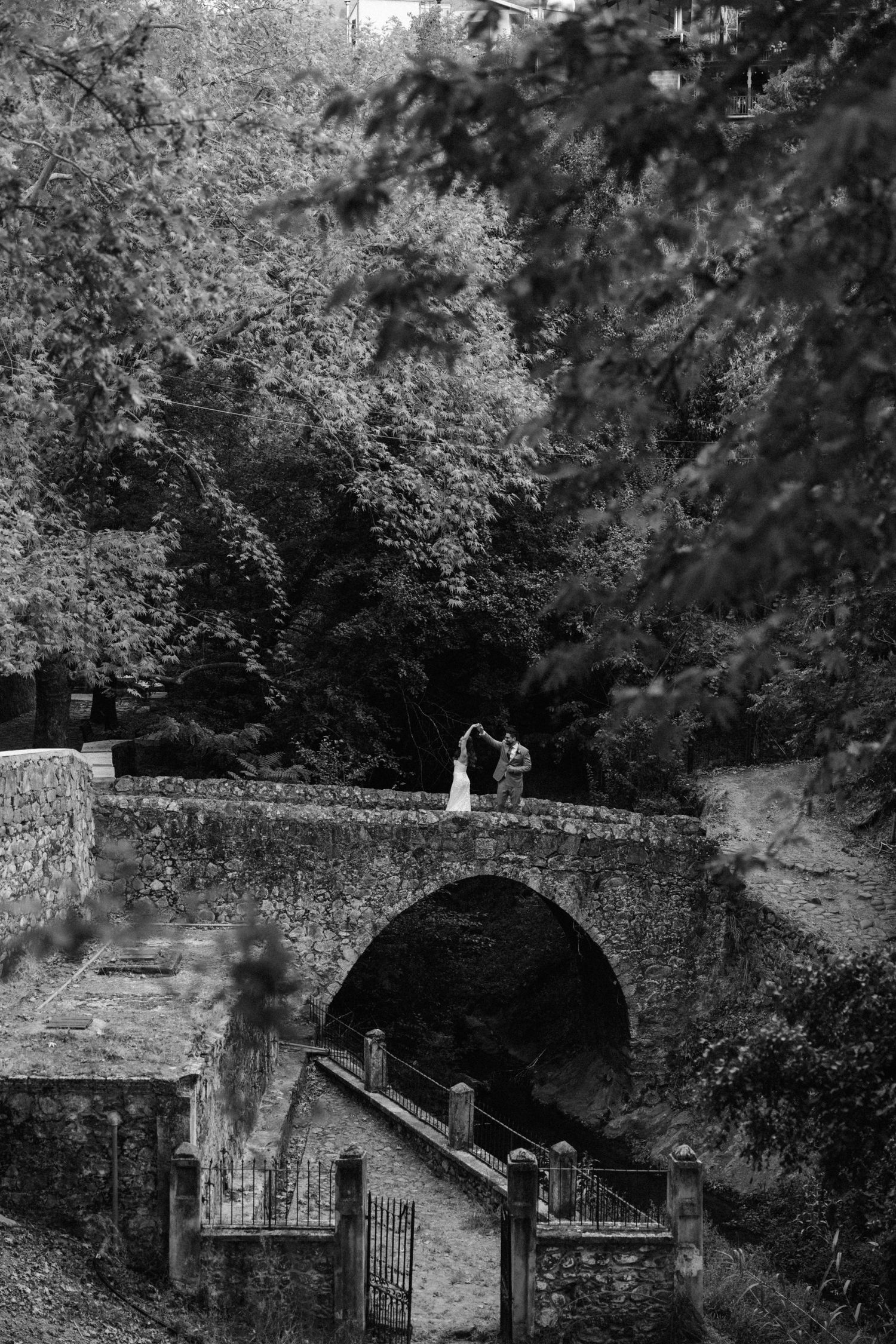 Black and white wide shot of couple on historic stone bridge surrounded by trees.