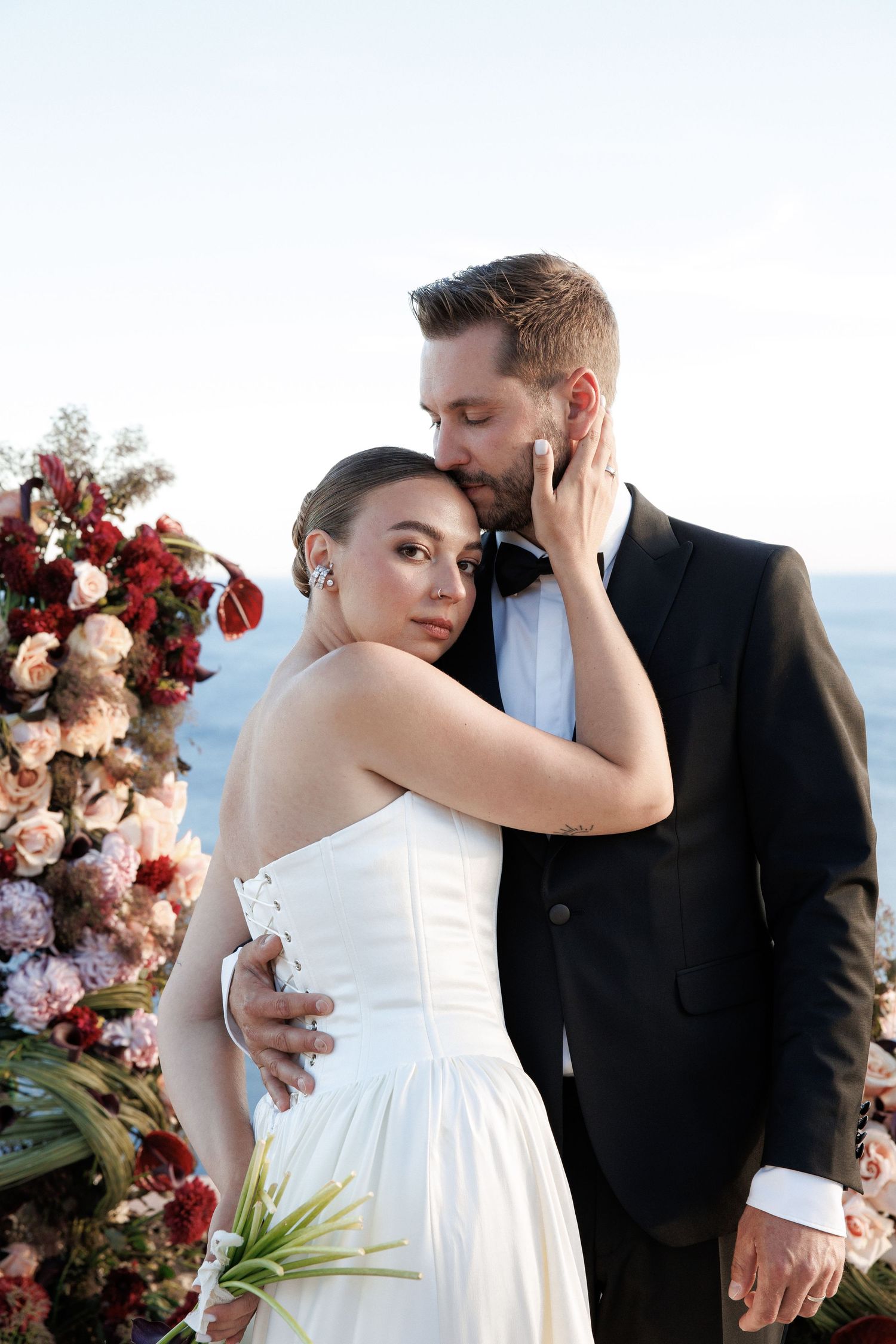 Groom tenderly embraces bride beside floral arrangement at cliffside wedding ceremony overlooking the sea.