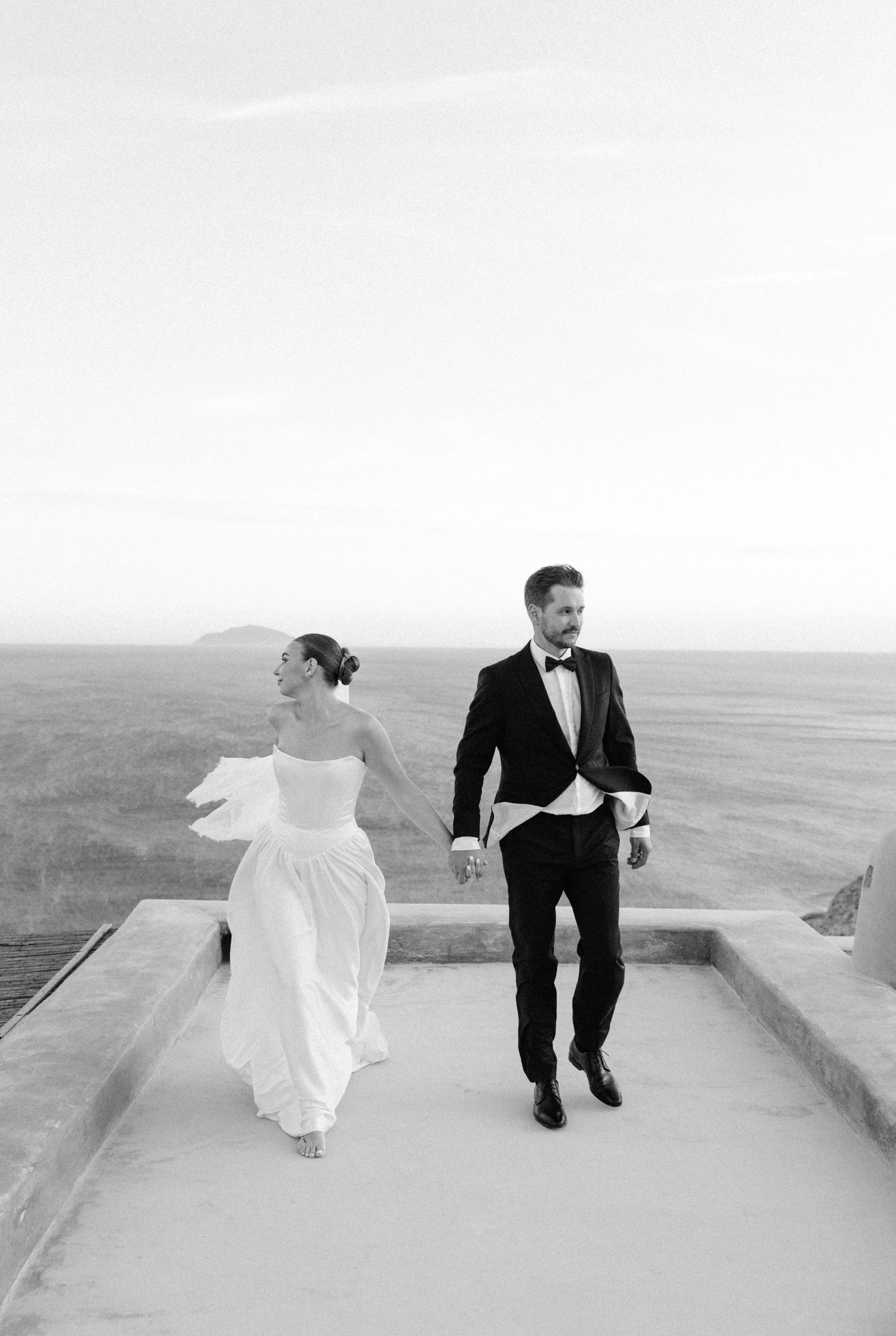 Black and white photo of bride and groom holding hands walking on rooftop terrace with ocean view.