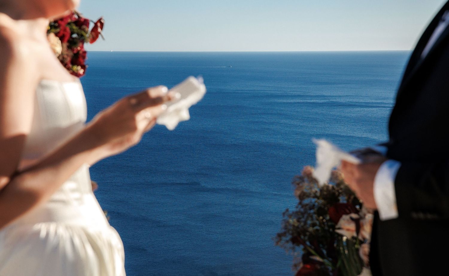 Bride in white dress reading vows by the ocean at a cliffside wedding ceremony with blue sea backdrop.