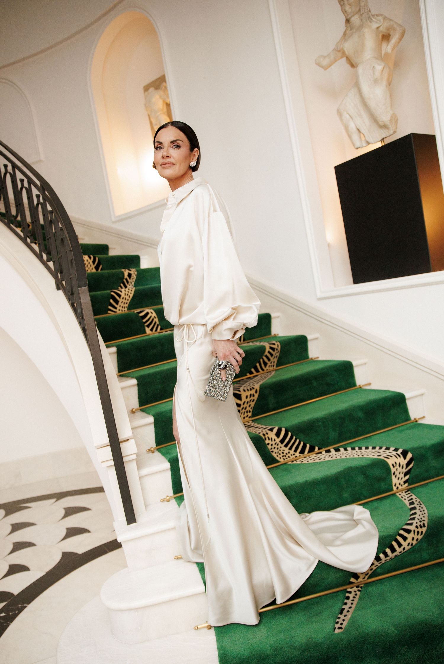 Woman in elegant white satin gown poses on green velvet staircase in luxury interior with ornate gold railing.