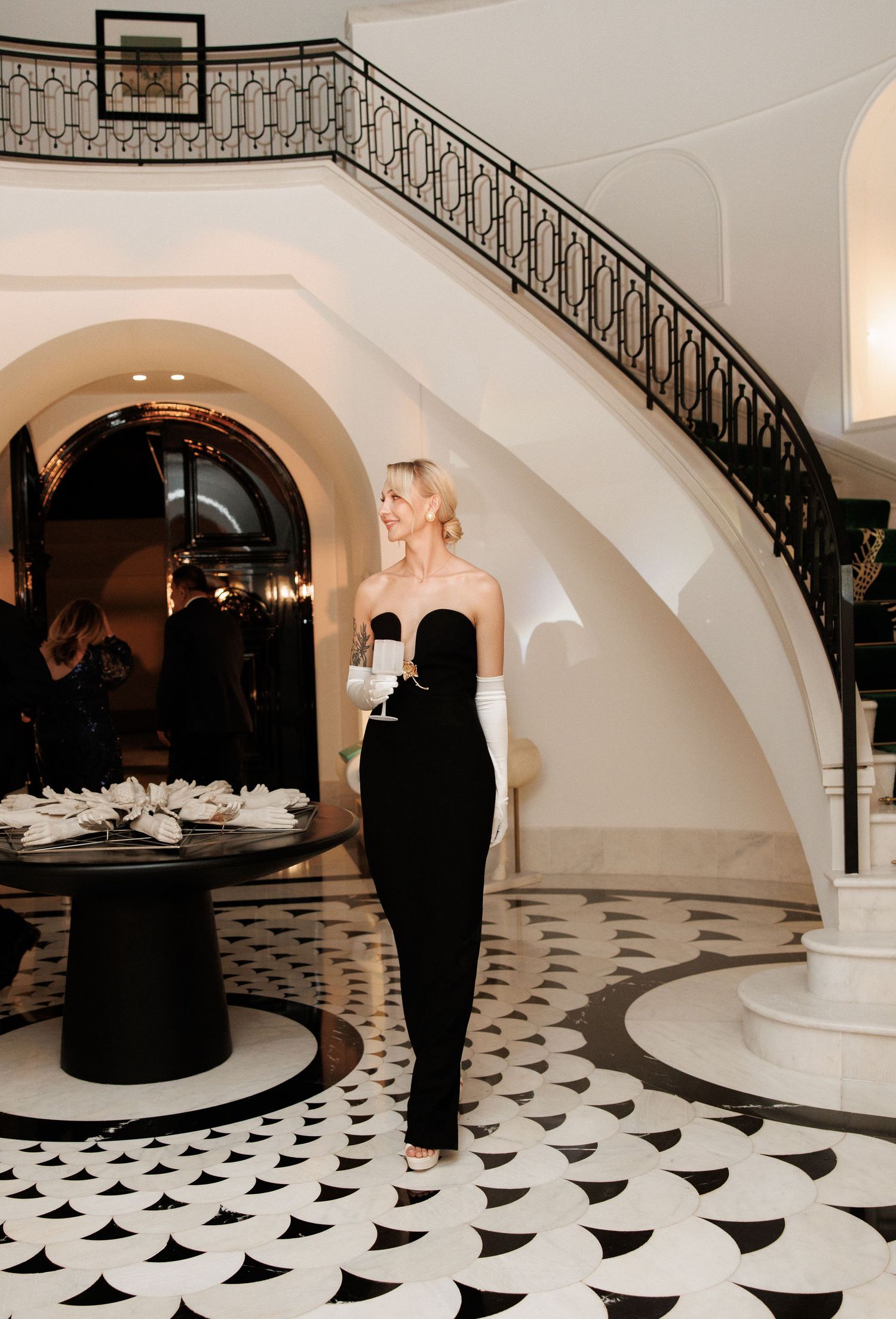 Elegant woman in black strapless gown holding champagne in luxury hotel lobby with grand staircase.